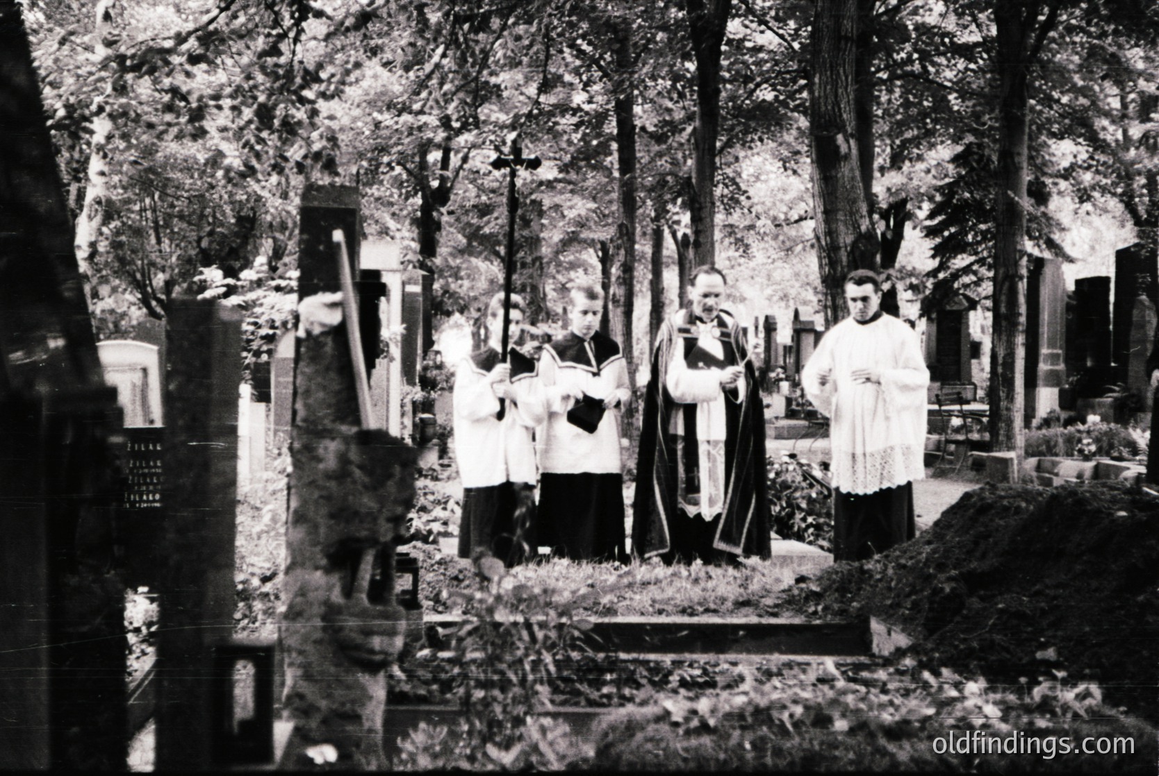 Mid-20th century black-and-white cemetery scene: clergy in liturgical vestments (cassocks, stoles) conduct a funeral service at a grave site surrounded by headstones. Lush forest backdrop suggests a rural or suburban setting.