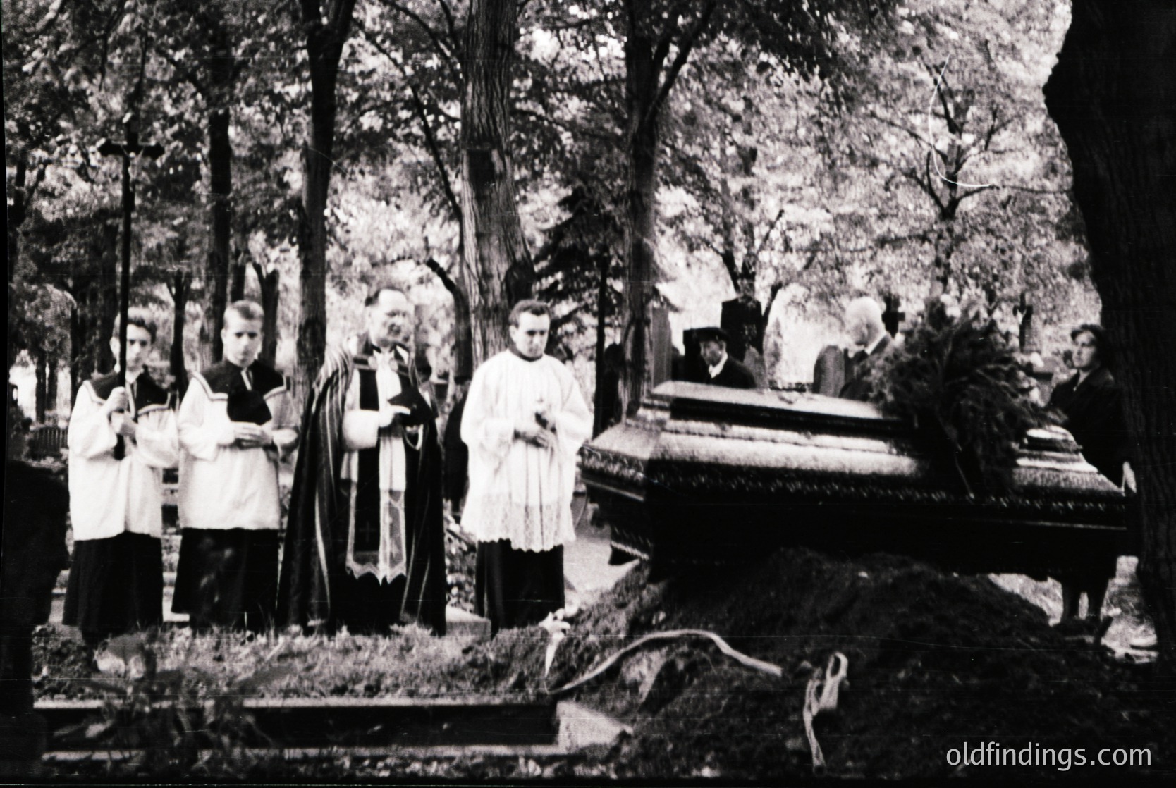 Black-and-white funeral procession in a forested cemetery, likely Eastern European . Clergy in liturgical vestments (cassocks, stoles) stand around an open grave, holding prayer books. Dense trees frame the scene, emphasizing solemnity. Grave markers and a draped coffin suggest a formal burial ritual.