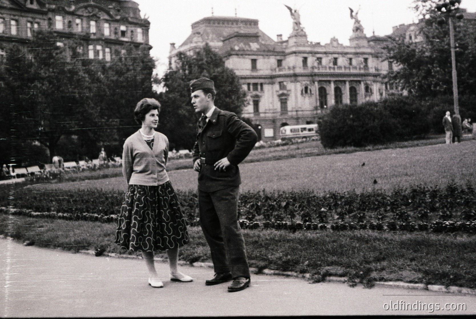 Mid-20th century black-and-white photo: two individuals in formal attire—man in military-style jacket, woman in patterned skirt and cardigan—standing on a landscaped park path. Ornate building with statues in background suggests European urban setting.