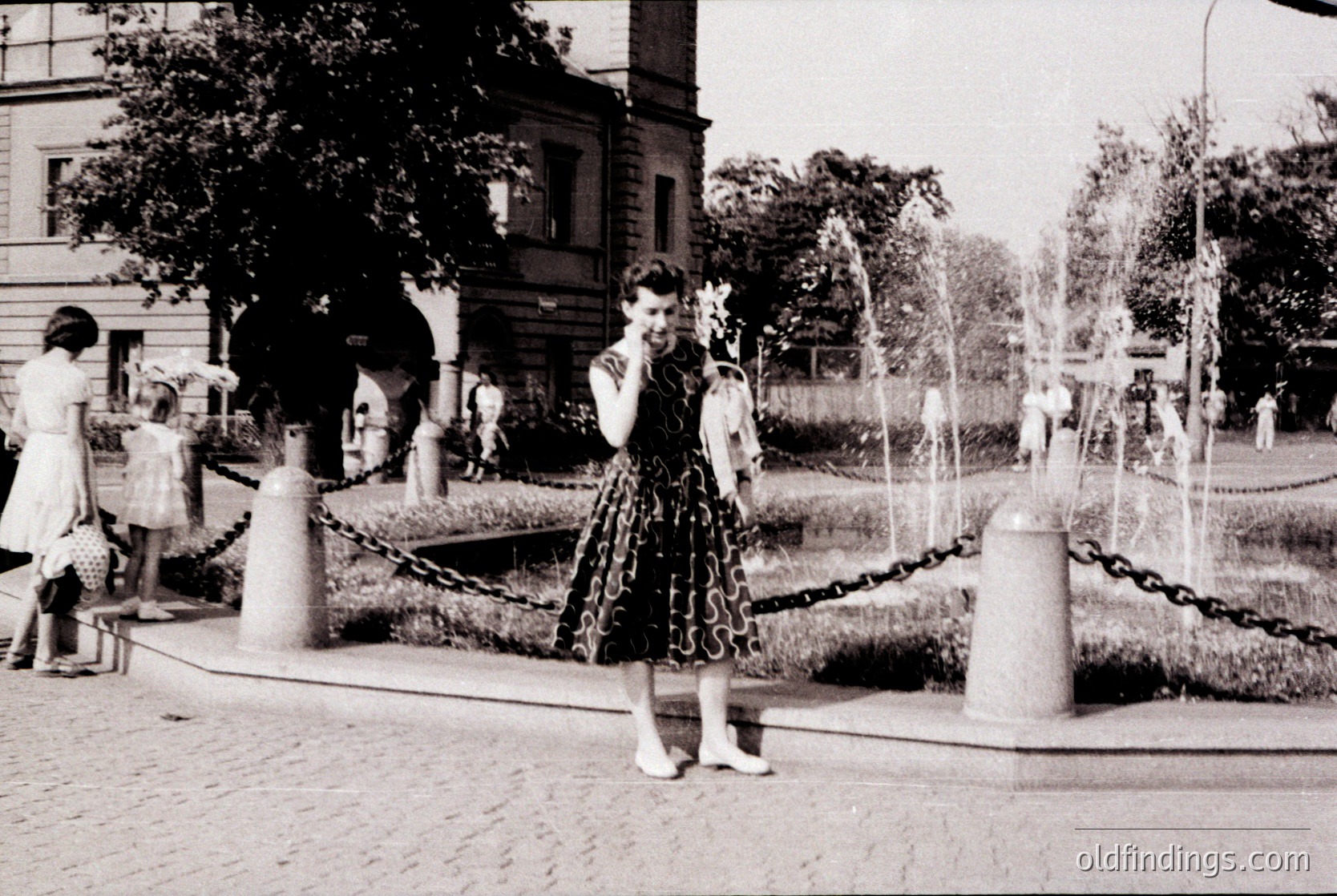 Vintage black-and-white photo of a woman in a 1950s-style dress with floral appliqué, posing near a fountain in an urban park. Chain barriers and decorative posts frame the scene. Mid-century architecture and greenery in background.