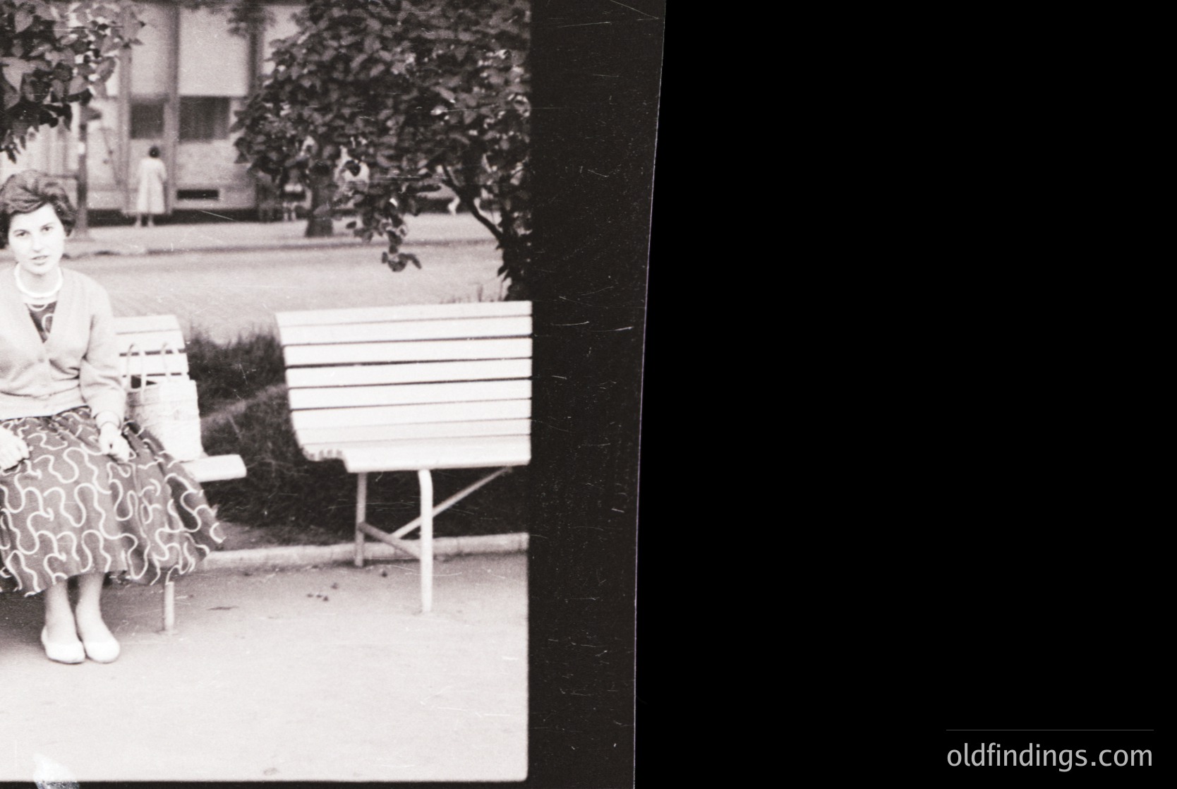 Mid-century woman in patterned dress sits on a simple wooden bench in a park-like setting. Classic 1950s–1960s fashion with structured skirt and cardigan. Urban greenery and vintage architecture in background.