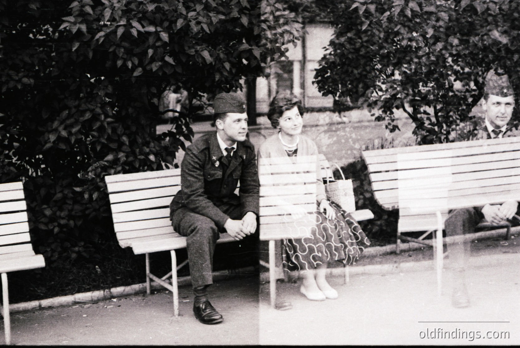 Vintage black-and-white photo of two individuals seated on park benches in a shaded area, likely mid-20th century. The man wears a military-style cap and jacket, while the woman’s patterned dress and hairstyle suggest 1950s fashion. Lush greenery and a blurred background suggest an urban park setting.