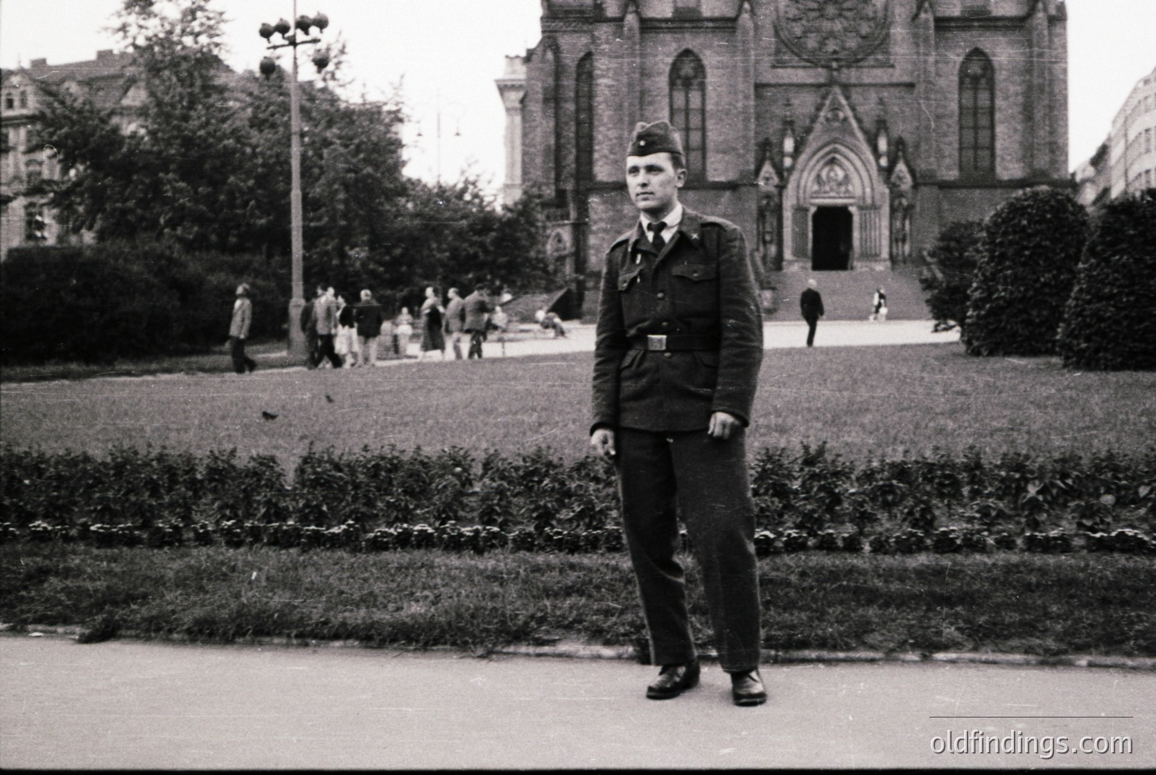 Mid-20th century uniformed man in a landscaped urban park, Gothic-style church with pointed arches in background. Black-and-white photo, likely 1950s–1960s.