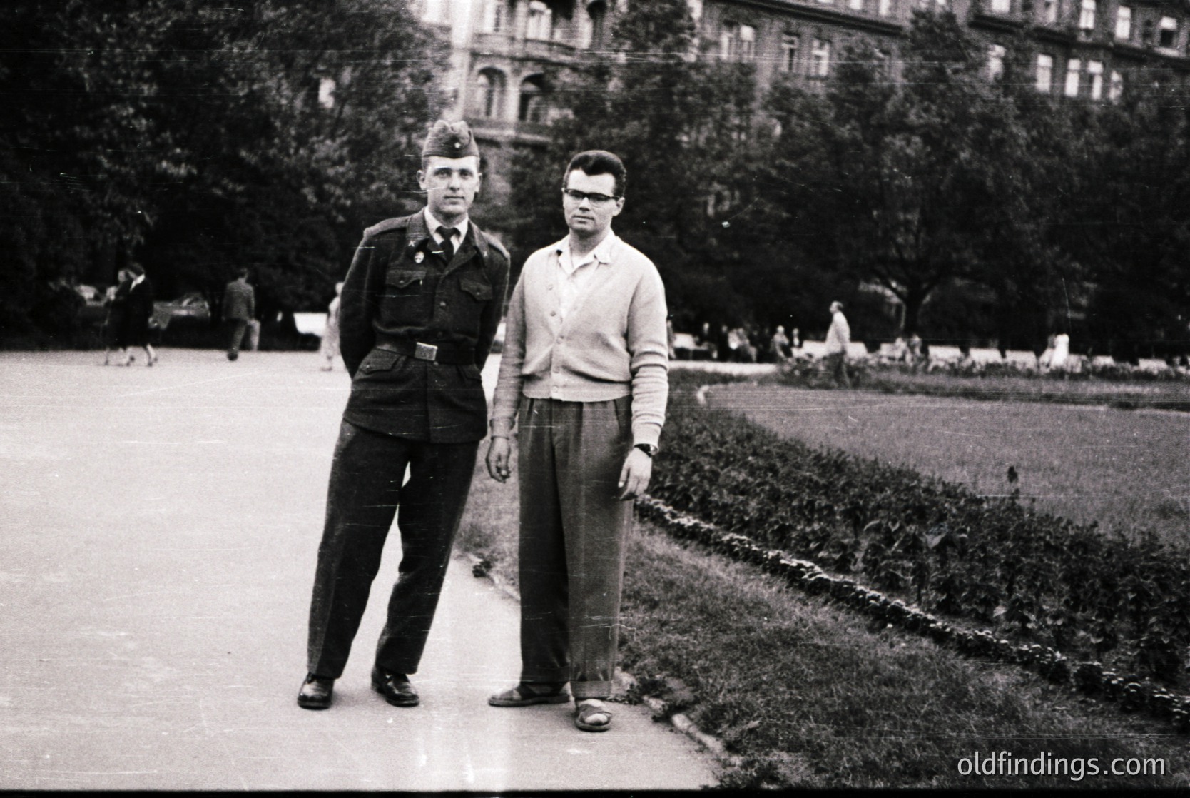 Black-and-white portrait of two men in 1950s European urban park. Left: uniformed individual in peaked cap, belted jacket, and trousers. Right: civilian in sweater, glasses, and rolled pants. Background features manicured gardens, classical architecture, and paved pathways.