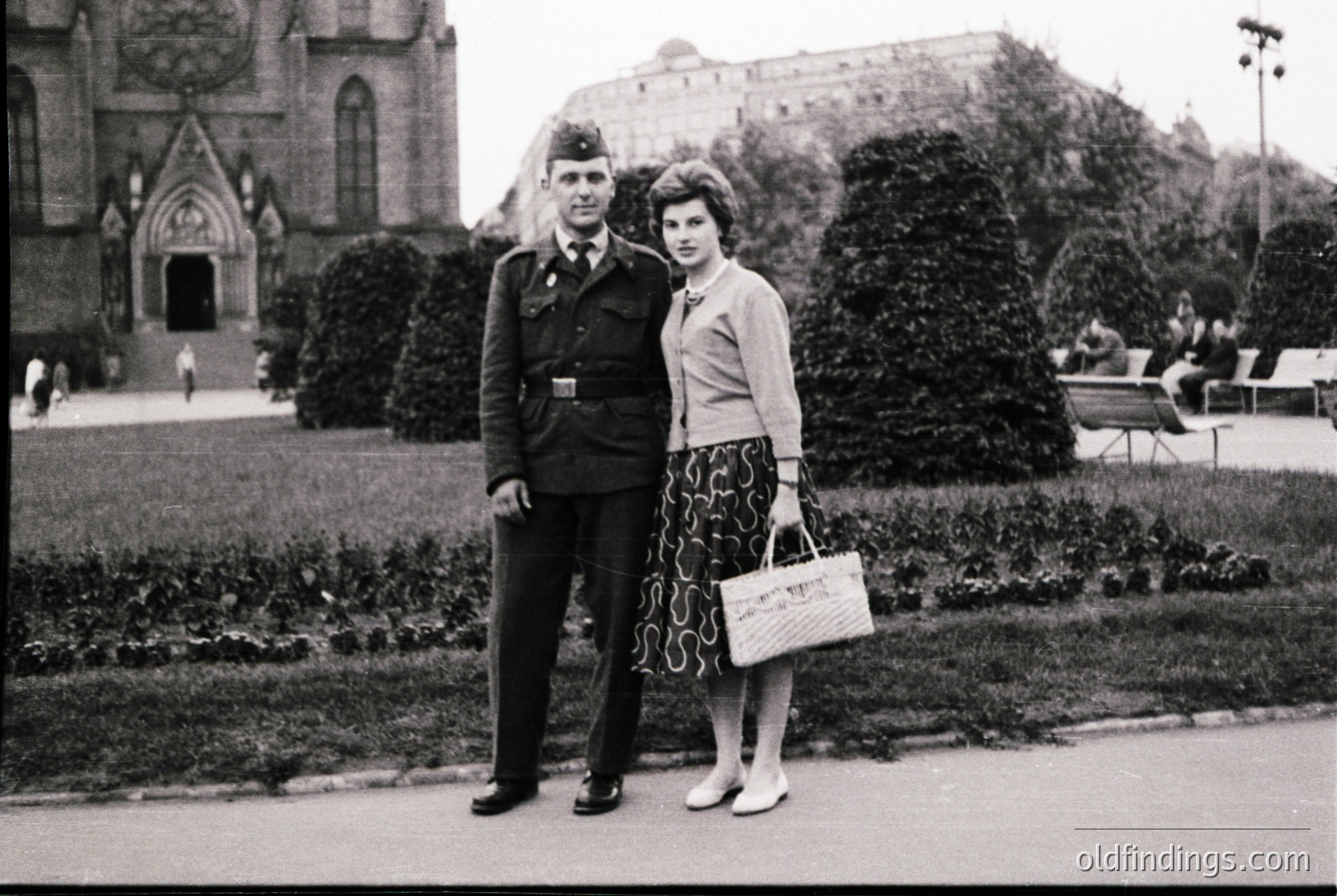 Man in 1950s military uniform (likely Bulgarian People’s Army) poses with woman in patterned skirt and cardigan in a landscaped park. Gothic-style church and Soviet-era building in background suggest Sofia, Bulgaria. Candid, mid-century urban portrait.