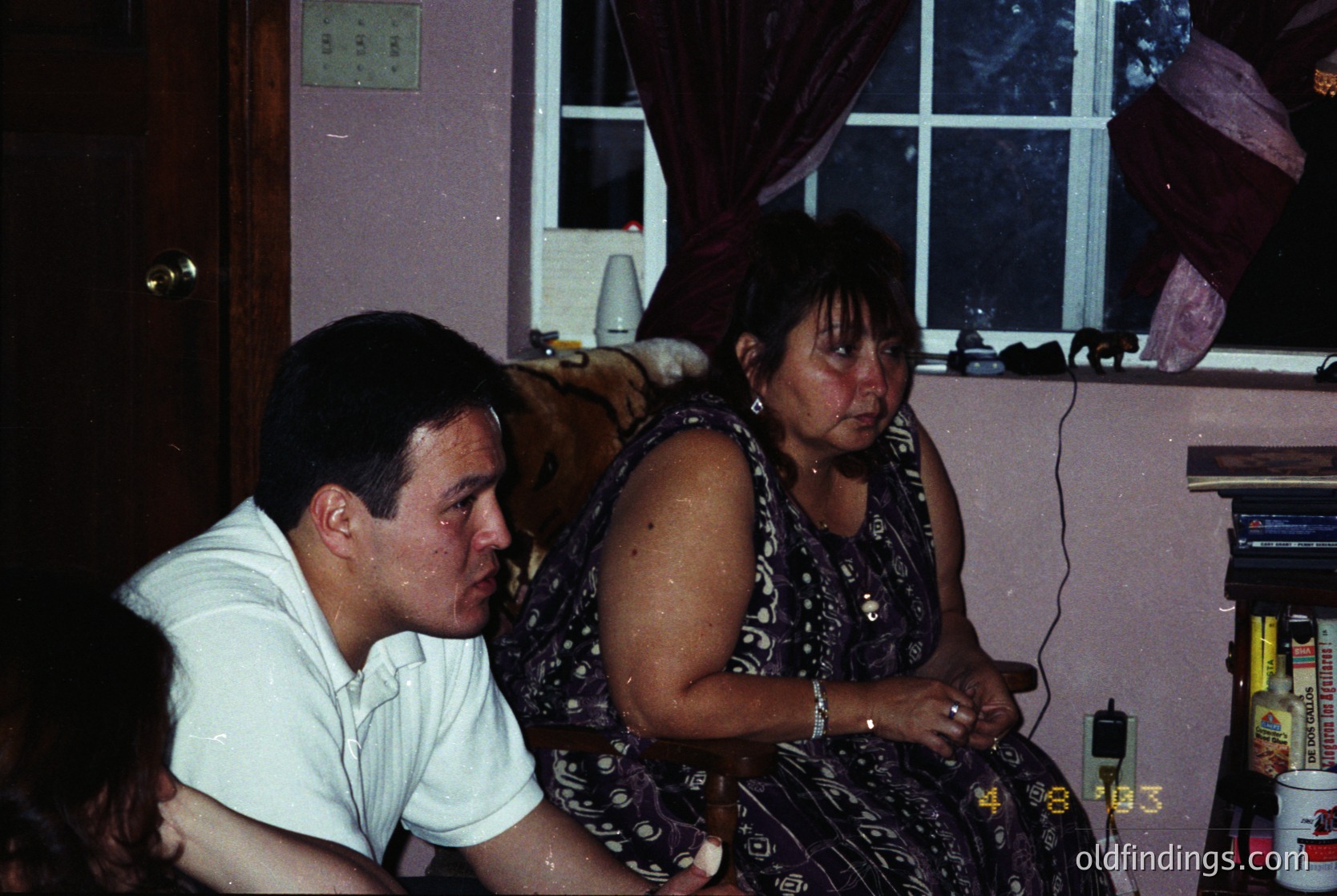 Vintage indoor portrait of two seated individuals in a dimly lit room, likely mid-20th century. The man wears a white button-down shirt, while the woman’s patterned blouse and skirt suggest 1970s–1980s fashion. A dog rests on the woman’s lap, and a shelf behind them holds decorative items and a radio. Warm, grainy film captures a candid, intimate moment.