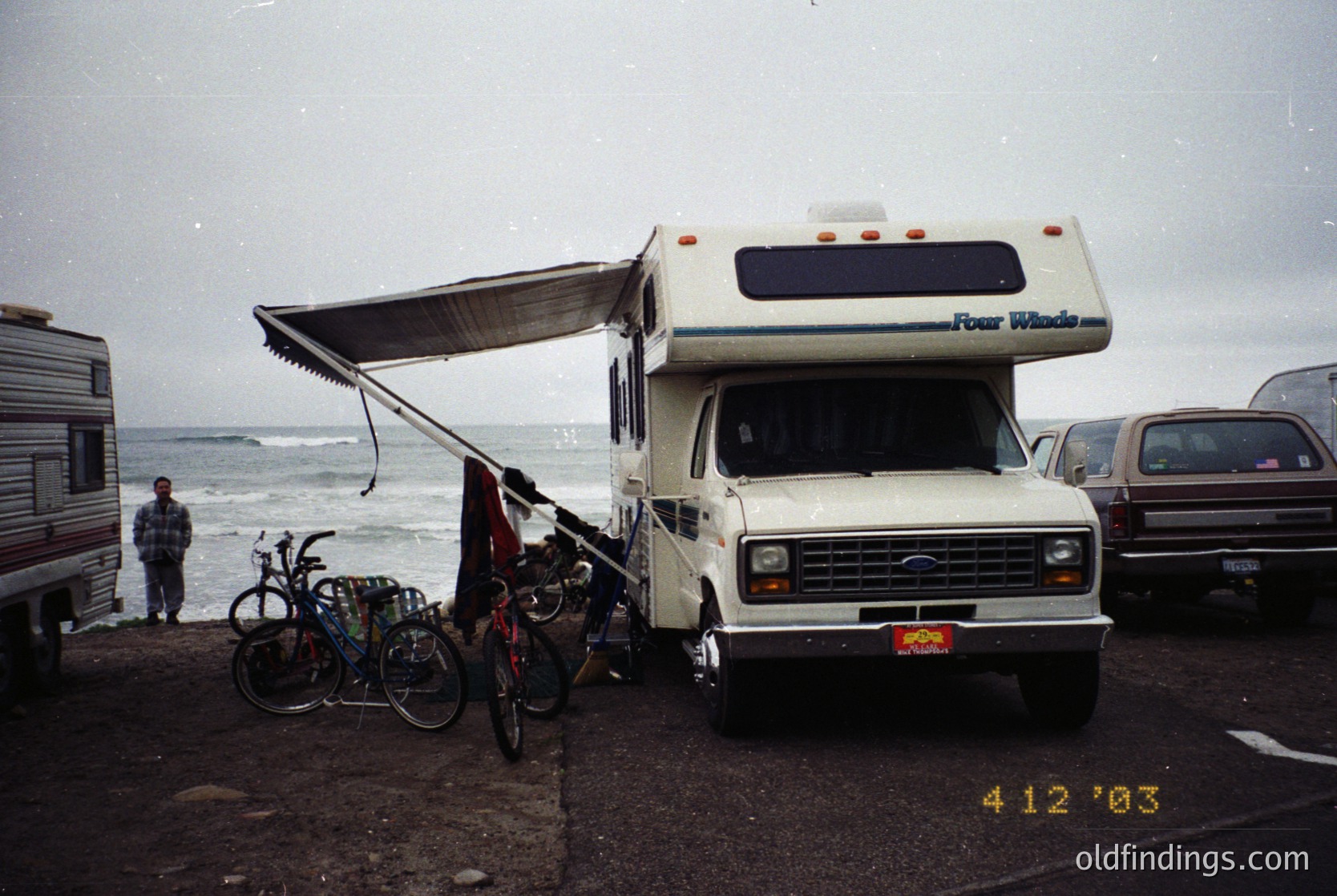 Classic 1980s-era Four Winds RV parked by a coastal road, with extended awning. Two bicycles secured to the front, one with a child seat. Overcast seaside setting with waves visible. Timestamp suggests early 2000s ().