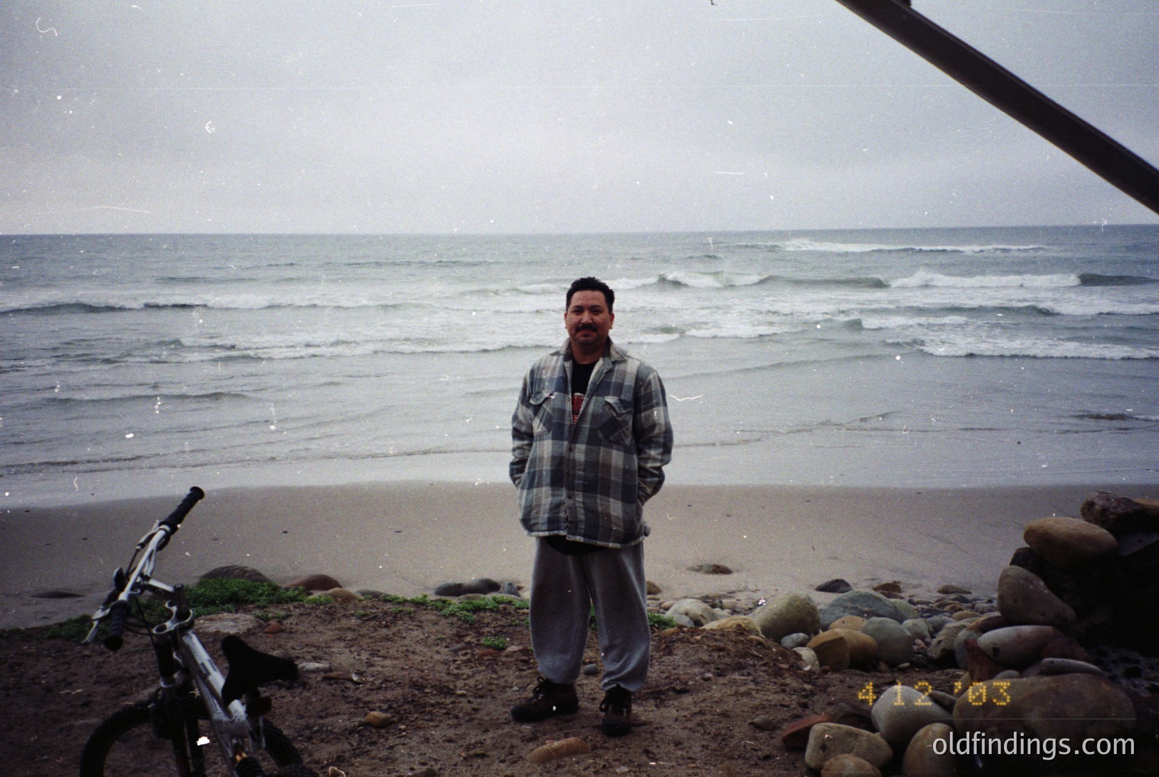 Man in plaid shirt and dark pants stands on a rocky seaside beach, rain-soaked waves crashing behind. Black bicycle leans against a metal structure. Vintage digital camera watermark (2000s era). Coastal