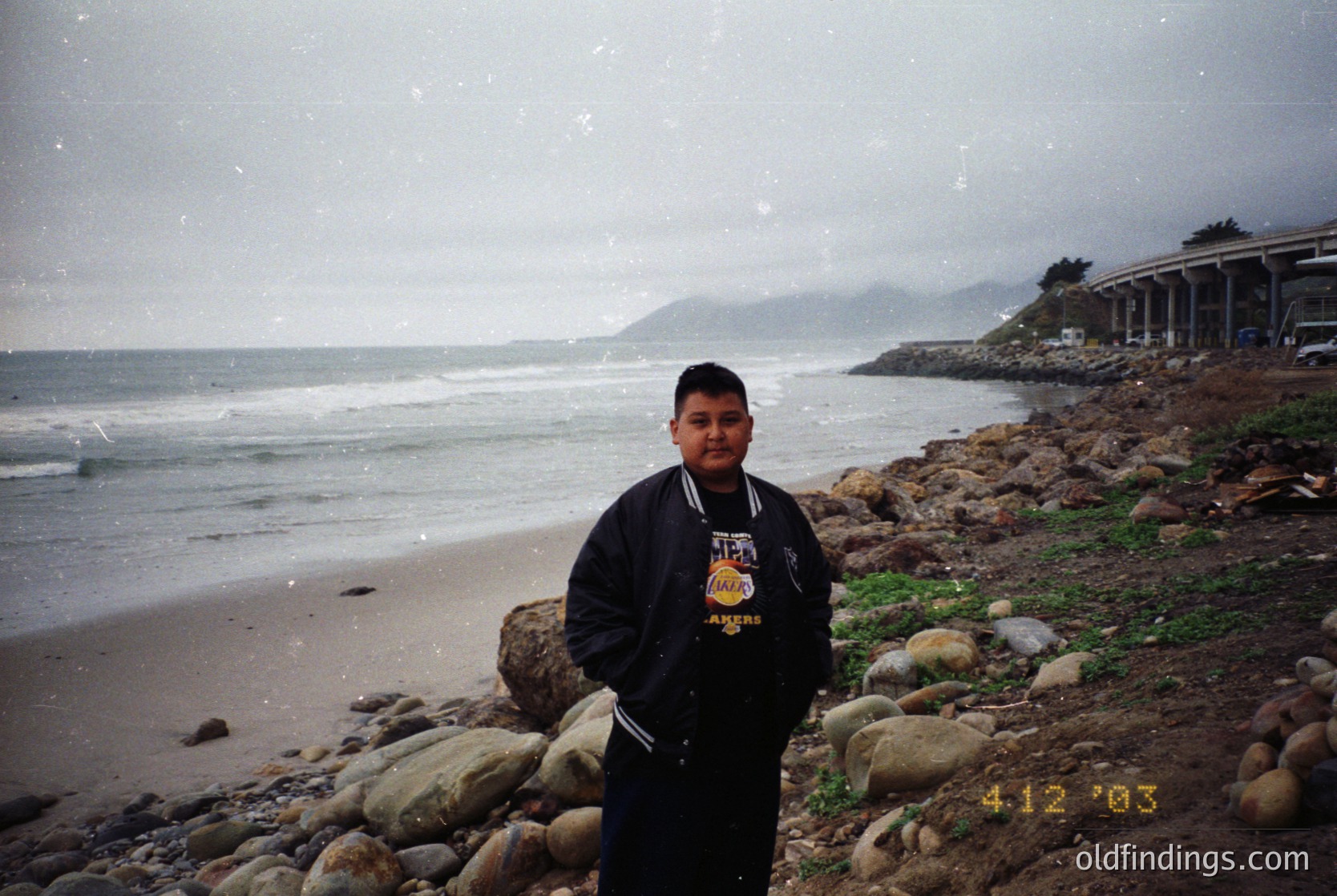 Young man in a black jacket with "AMER" embroidered on the back poses on a rocky beach during light snowfall, with coastal cliffs and ocean waves in the background. Date stamp suggests early 2000s.