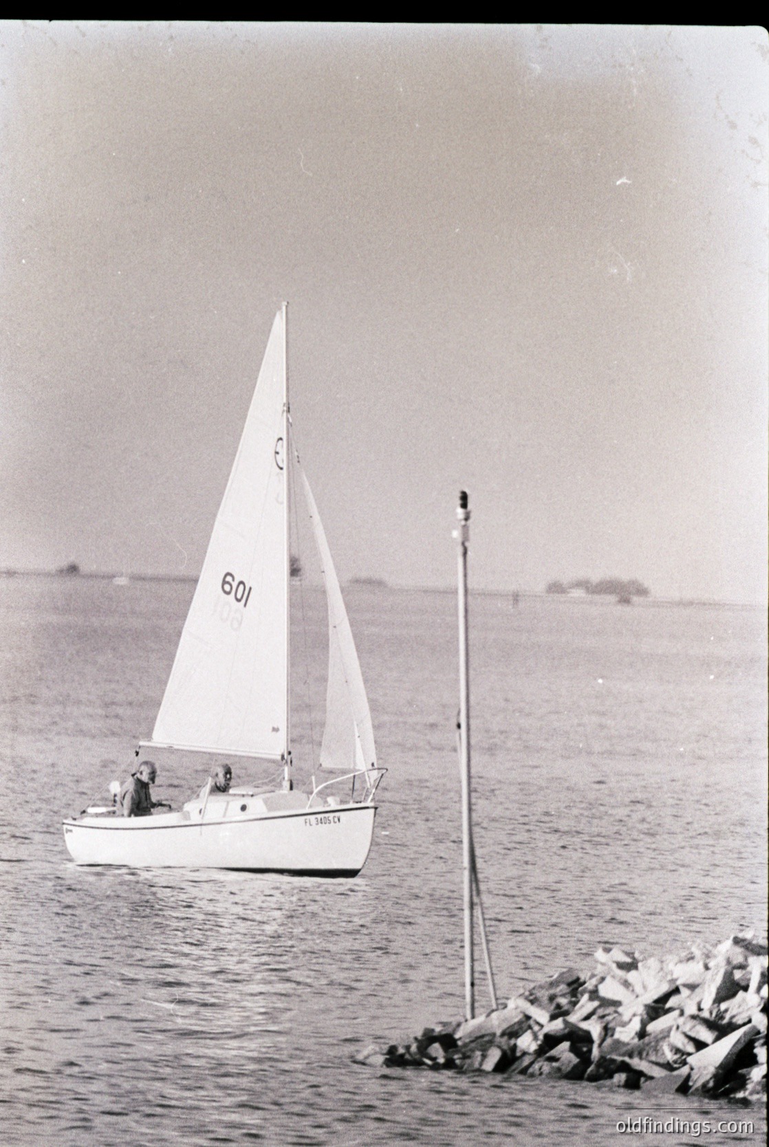 Vintage monochrome sailboat race photo, 1960s-70s. Small fiberglass dinghy () with two crew members, sailing near a rocky breakwater. Classic design with numbered sail and "LASER" hull label. Coastal waters, likely Mediterranean or Atlantic. Ideal for vintage sports, maritime history, or stock photography.