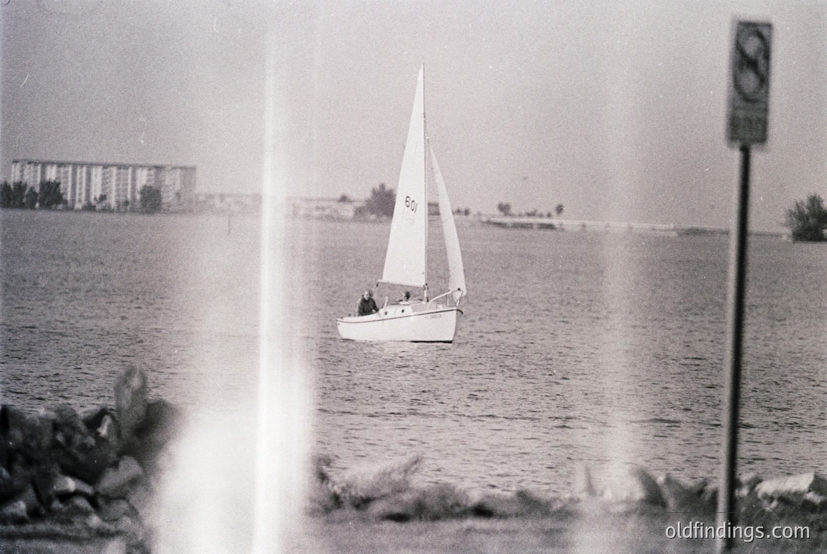 Vintage black-and-white sailboat on calm waters, framed by window or glass. Mid-20th century coastal architecture in background.