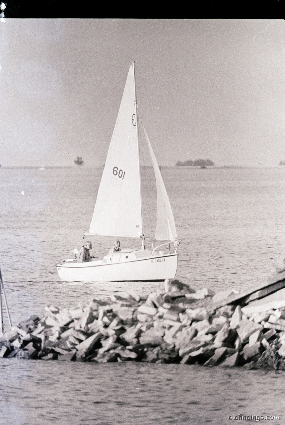 Classic 1960s-70s sailboat anchored near rocky shore, likely Mediterranean or coastal Europe. Minimalist design with single mast and white sail. Stormy seas suggest challenging sailing conditions. Ideal for vintage maritime archives or nautical design references.