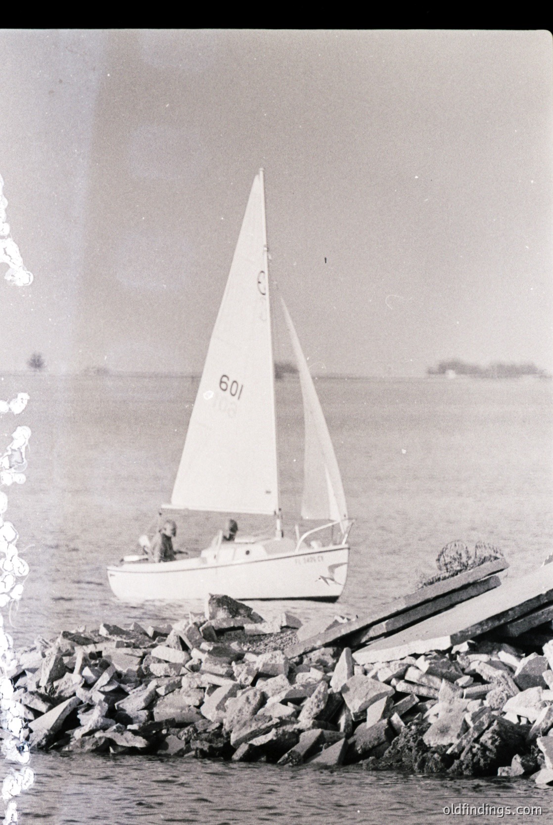 Vintage monochrome photo of a small sailboat () anchored near rocky breakwaters, likely mid-20th century (). Three figures visible on deck; distant shoreline and horizon suggest coastal setting. Ideal for maritime history or vintage travel references.