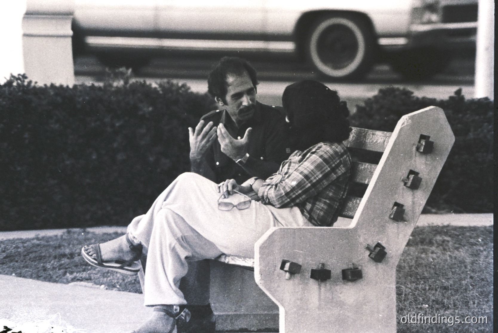 Two men in mid-20th-century attire share a moment on a modernist concrete bench, likely 1960s–1970s. The man on left wears light pants and a jacket; the other, a plaid shirt and sunglasses. Background features a blurred car and landscaped greenery, suggesting an urban park or campus setting.