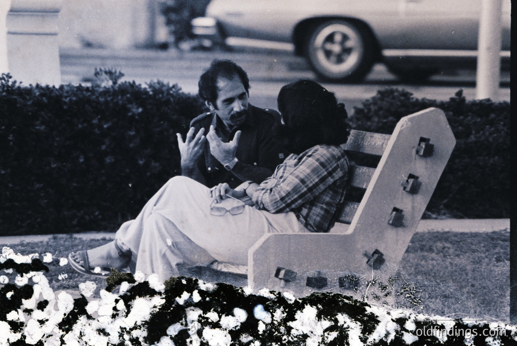 Black-and-white candid of two individuals seated on a modernist concrete bench in a landscaped park setting, 1960s–1970s. One wears a light-colored tunic, the other a plaid shirt. Blurred vintage car and manicured hedges in background suggest suburban or institutional location.