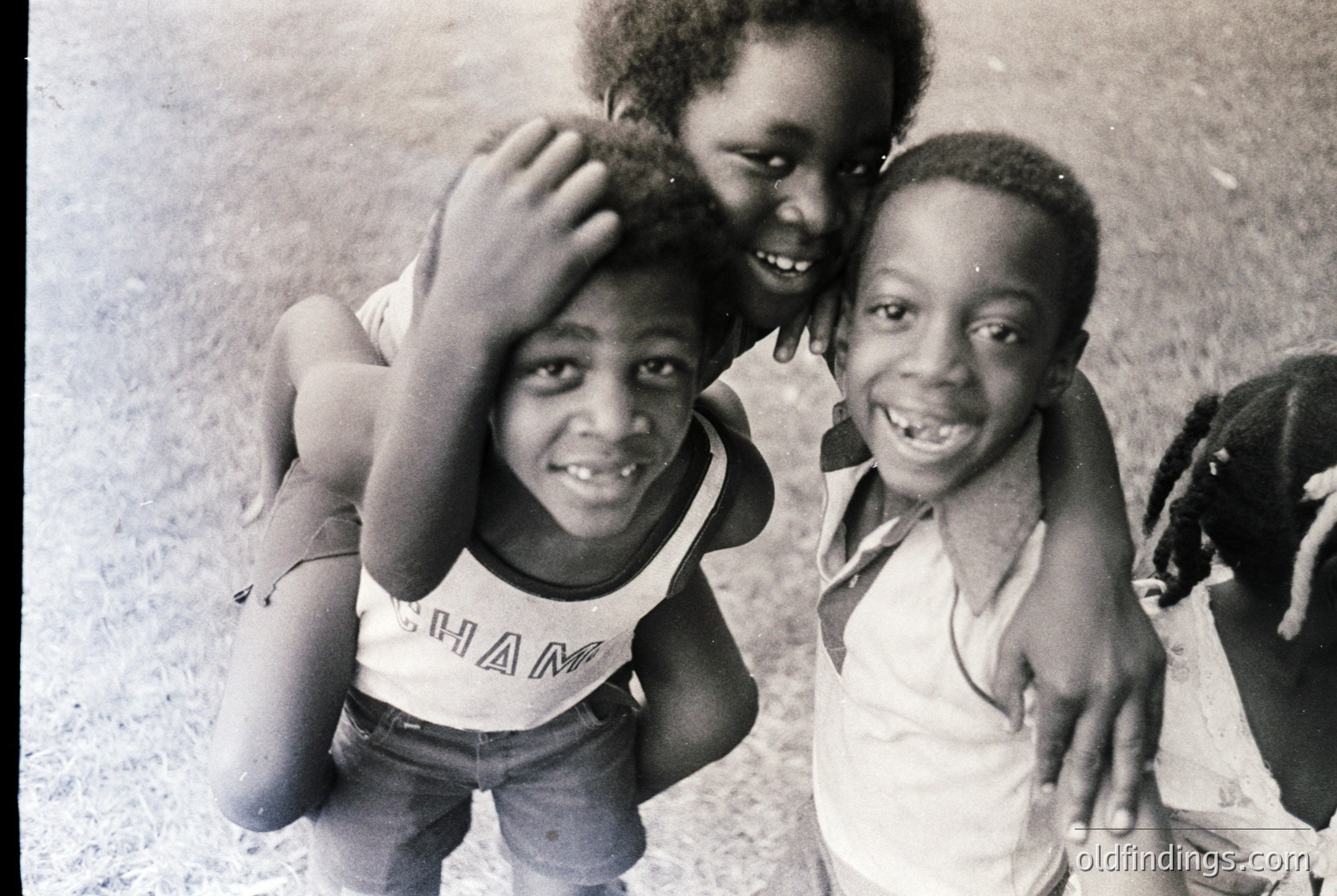 Black-and-white candid of three joyful children posing outdoors, likely mid-20th century. The boy in the center wears a "CHAM" tank top, while the others sport open-collar shirts. Urban setting with rough pavement and blurred background. Evokes 1960s–1970s street culture.