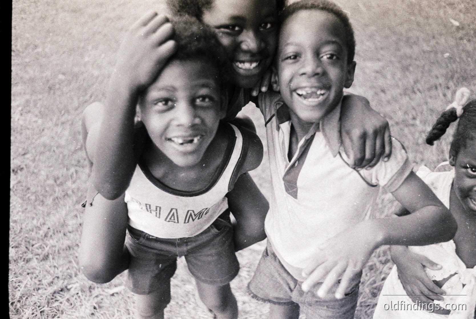 Black-and-white candid of three joyful boys in outdoor setting, mid-1960s–70s. Foreground boy wears "Champion" tank top and shorts; others in casual attire. Grass and blurred background suggest park or backyard. Authentic 20th-century childhood energy.