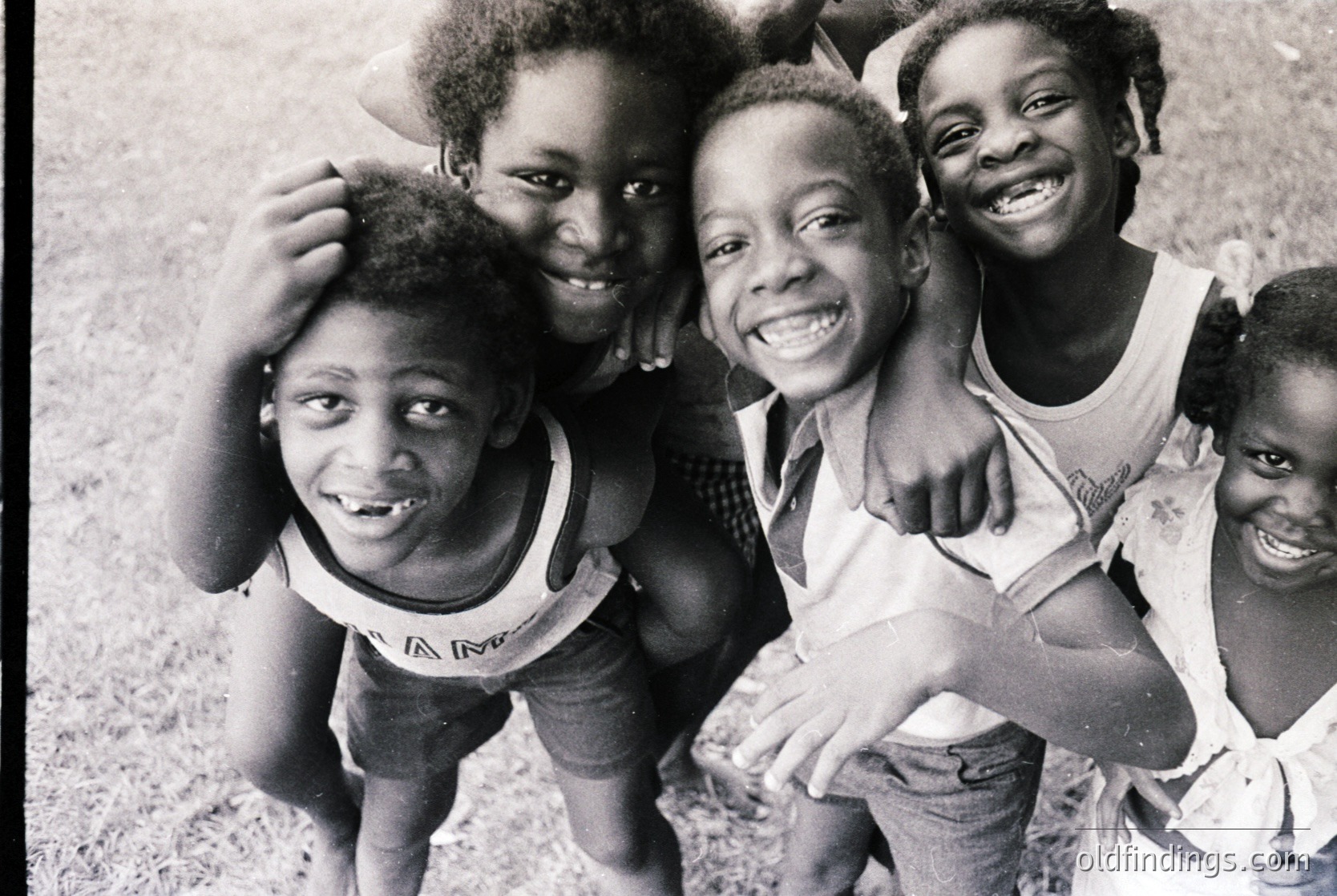 Four joyful children pose closely in a 1970s outdoor setting, wearing matching white tank tops with floral patterns. Their broad smiles and relaxed postures suggest a carefree moment. Likely captured in a community or school event.