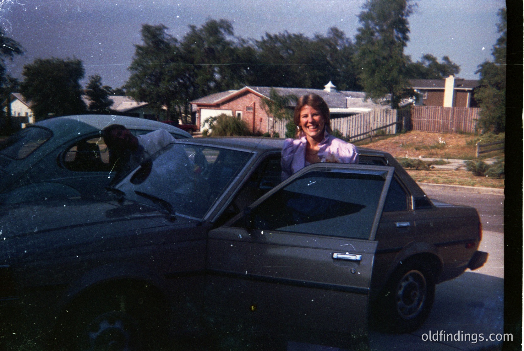 Vintage 1970s-era station wagon with a woman leaning out the driver’s side, smiling. Suburban residential setting with brick houses, palm trees, and a gated backyard. Film grain and sepia tone enhance nostalgic feel.