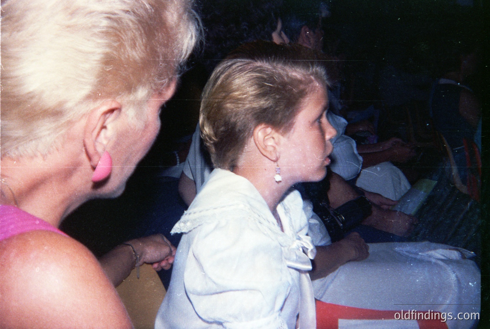 Vintage candid shot of two women in close embrace, likely at an event. The elder woman wears a pink sleeveless top, while the younger, in a white blouse with a bow, appears emotional. Blurred crowd and indistinct red signage suggest a public gathering or ceremony. Style and lighting hint at 1970s–1980s indoor photography.