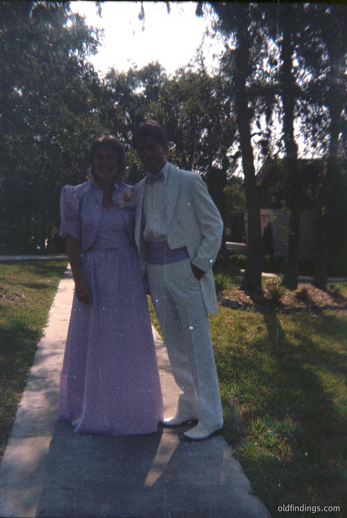 Couple in formal attire—woman in a fitted purple gown with a high collar, man in a light-colored suit with a sash—posing outdoors on a paved path surrounded by trees. Likely a 1950s–1960s wedding or formal event.