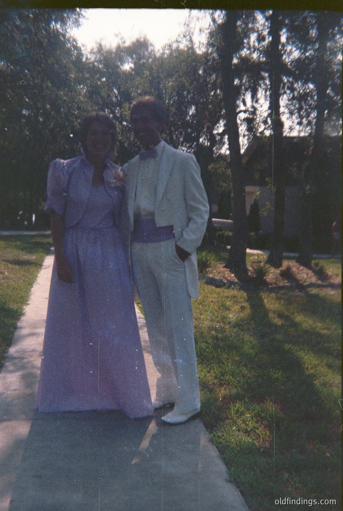 Couple in formal attire—woman in a knee-length purple dress with a belt, man in a light-colored suit with a sash—posing outdoors on a paved path surrounded by trees. Likely 1950s–1960s wedding or formal event.