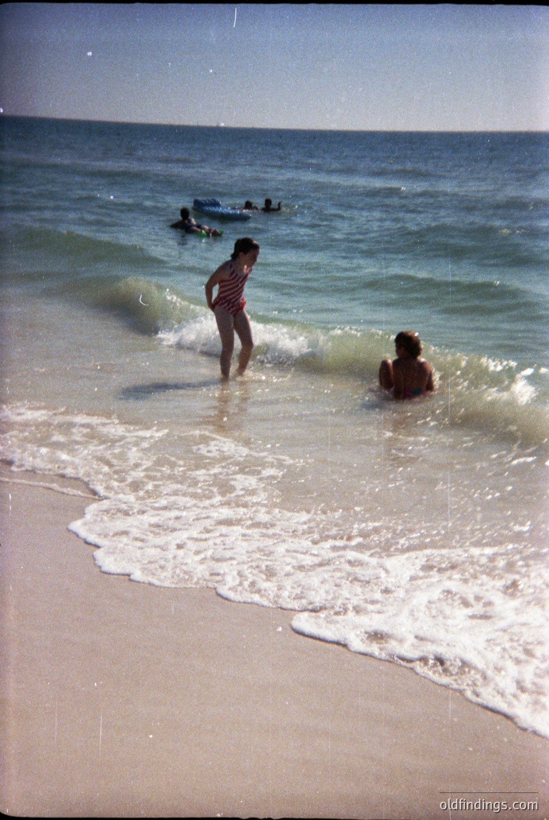 Vintage seaside scene with two children wading in shallow waves, one standing in a striped shirt, the other kneeling in swimwear. Gentle surf meets sandy shore under a clear sky. Likely mid-20th century beach culture.
