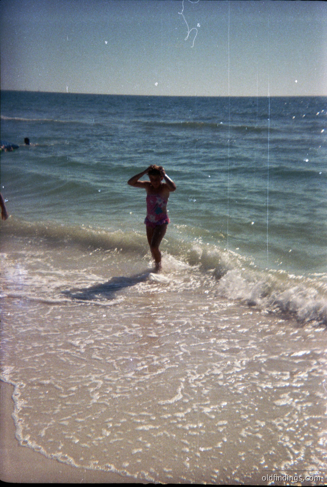 Vintage seaside scene: Woman in 1960s-style one-piece swimsuit wading into shallow waves, hand shielding eyes from sun. Light-colored sand and clear horizon suggest a Mediterranean or Atlantic coast. Film grain and slight overexposure indicate mid-century photography.
