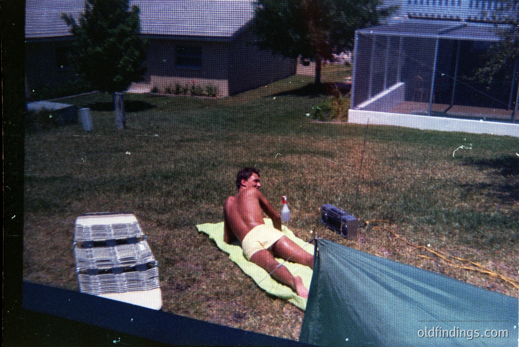 Vintage backyard scene featuring a shirtless man reclining on a green towel, holding a plastic bottle. Surrounding elements include a white plastic chair, a blue cooler, and a green mesh fence. Residential house with a metal roof and a birdcage in the background. Likely mid-20th century American suburban setting.