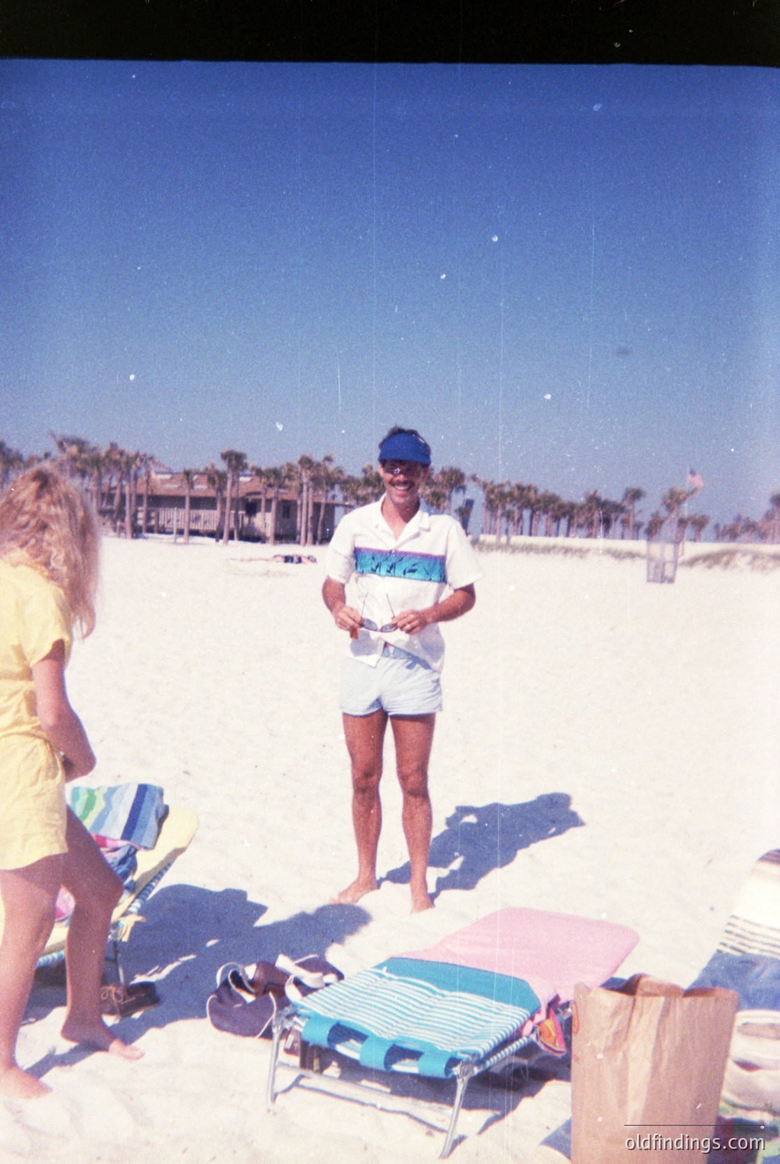 Vintage beach scene with two figures on sandy shore, palm trees in background. Man in 1970s swimwear (blue cap, striped shirt) poses with a camera, while woman in yellow top stands nearby. Folded beach chair and towel bag suggest midday relaxation. Clear skies and bright lighting indicate sunny coastal setting.