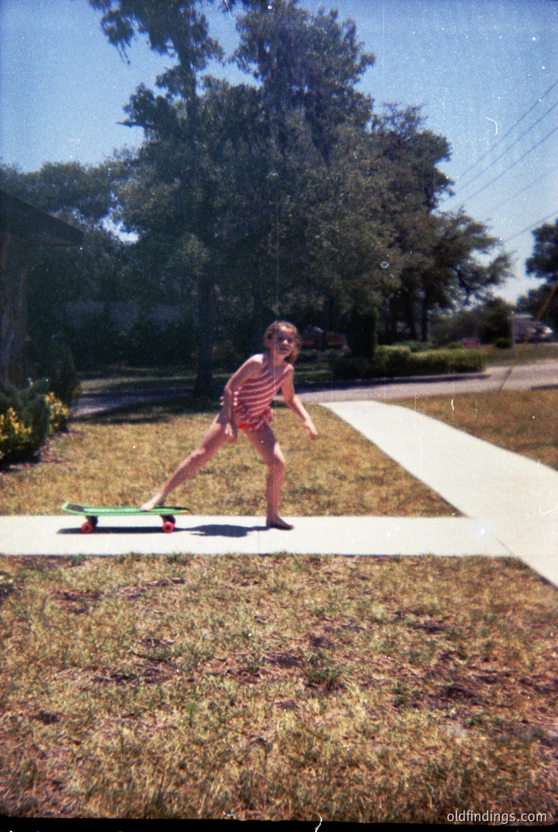 Vintage photo of a child skateboarding on a residential street, 1970s–1980s. Red-and-white striped shirt, barefoot, on a green skateboard with white wheels. Suburban neighborhood with dry grass, trees, and a curved driveway.