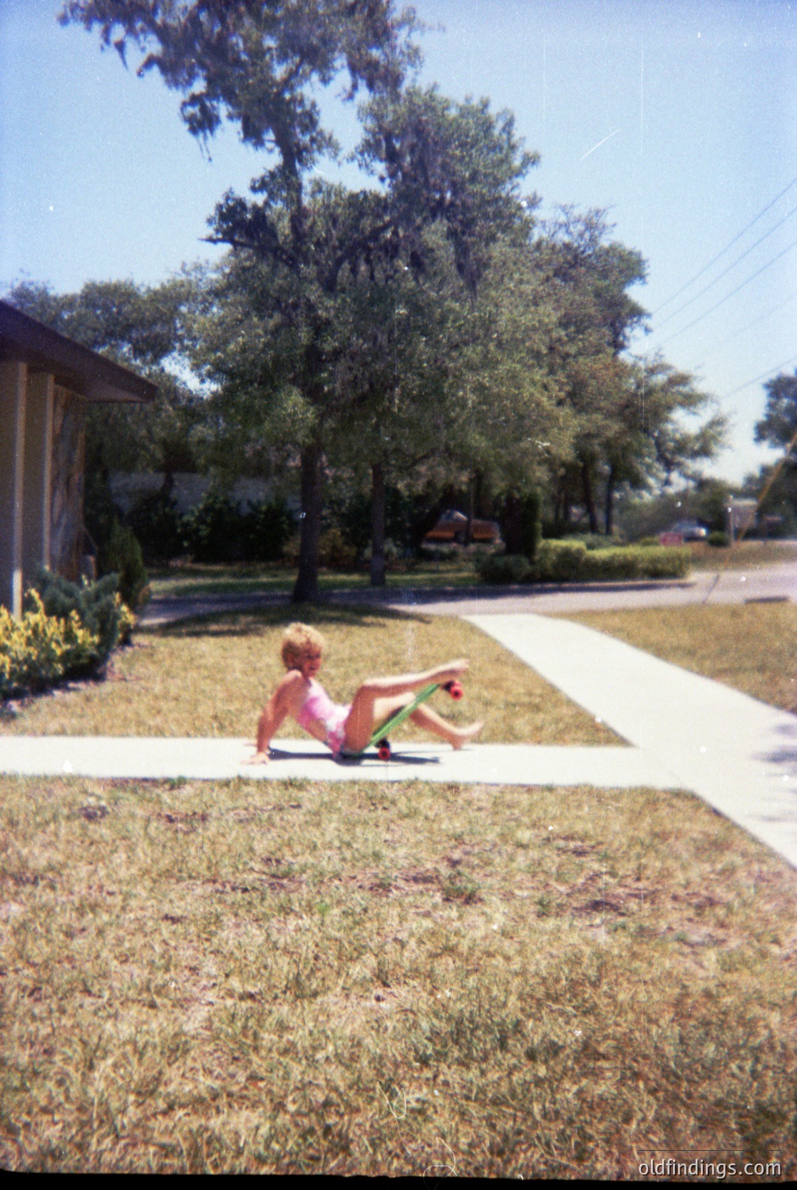 Child in mid-air skateboard trick on suburban sidewalk, 1990s-era residential street. Lush green trees and manicured lawns suggest suburban America. Classic skateboarding culture with vintage board design.