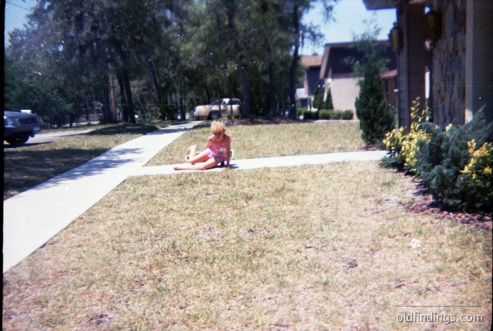 Child in 1970s-era suburban lawn, sitting on patchy grass near curb. Mid-century split-level home with dark trim and shrubs in background. Vintage Polaroid-style color saturation.