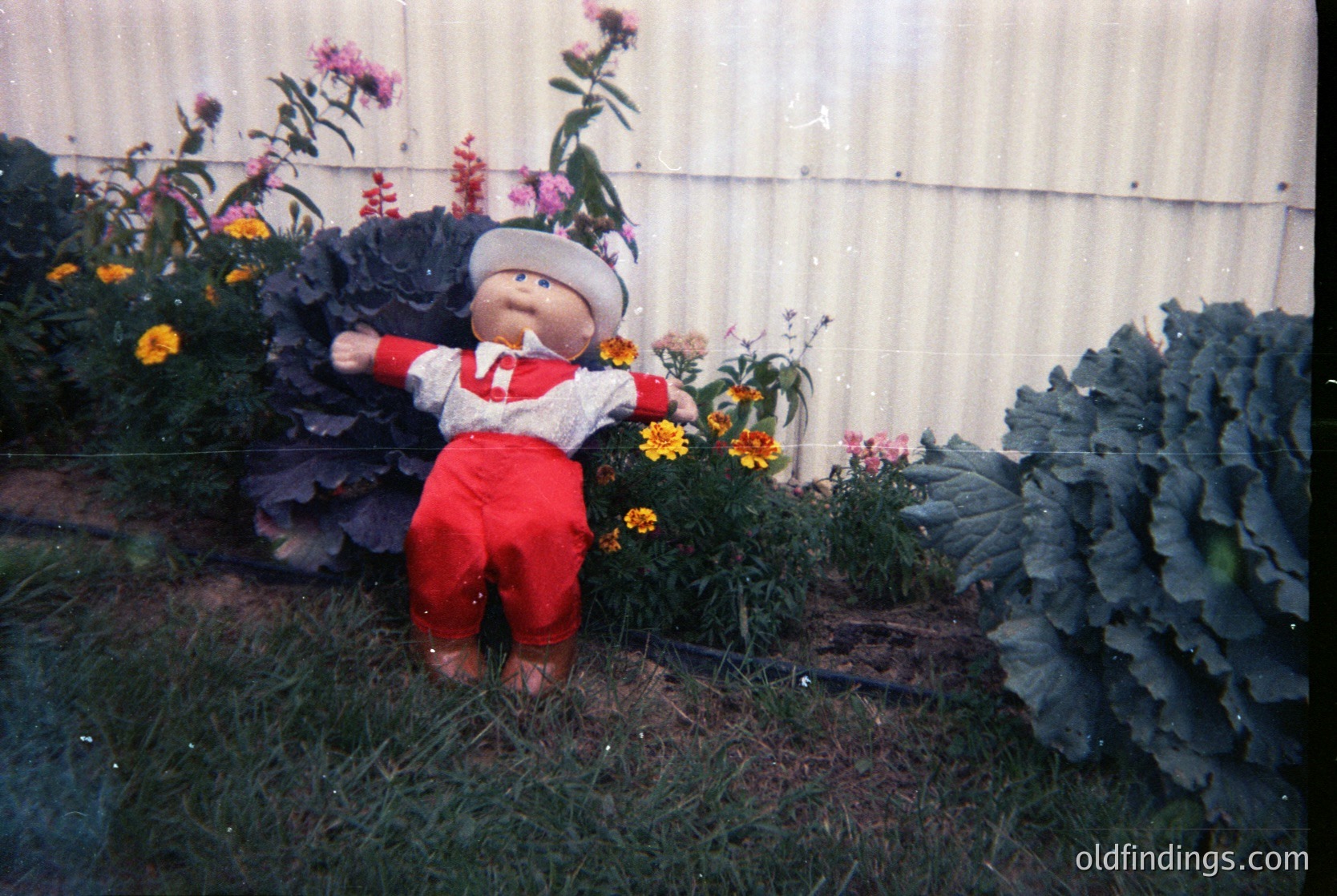 Vintage scarecrow-style doll in red overalls and white sweater, positioned among lush greenery and blooming flowers. The setting appears to be a garden or farm plot, likely from the mid-20th century. The doll’s design suggests a playful or decorative purpose.