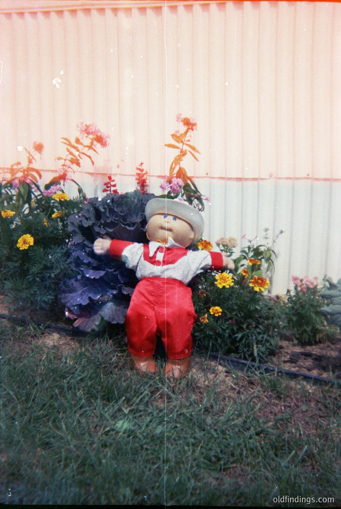 Vintage garden gnome figurine in red overalls and white apron, posed with arms outstretched among vibrant flowers and greenery. Likely mid-20th century (1950s–1970s) garden ornament.