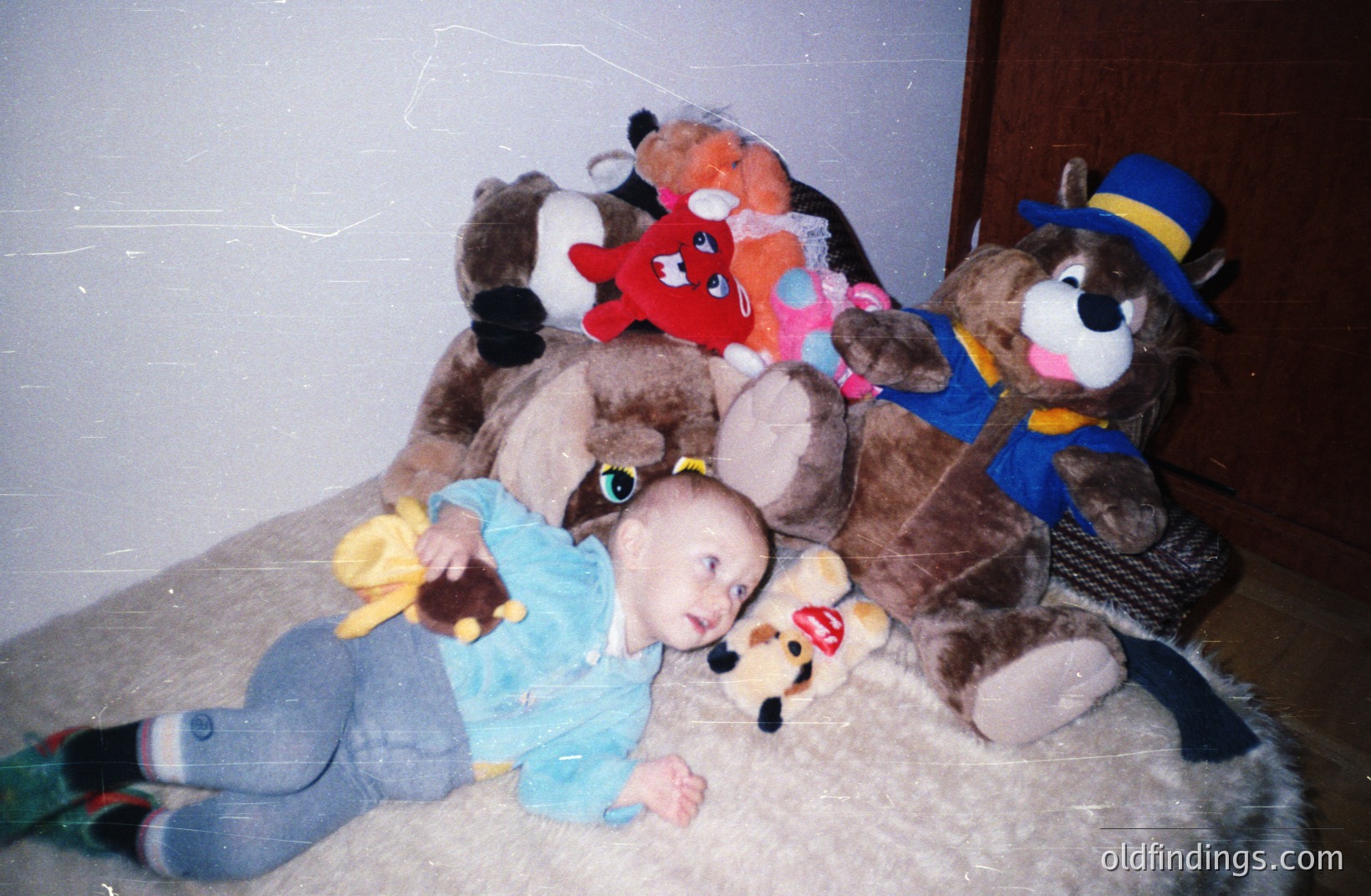 A young baby, clad in a blue shirt and leggings, reclines on a patterned rug, surrounded by several large, plush teddy bears and animal toys. The image's color palette suggests a snapshot likely taken in the 1970s or 1980s. A noticeable film grain & light leak reinforce the vintage feel.