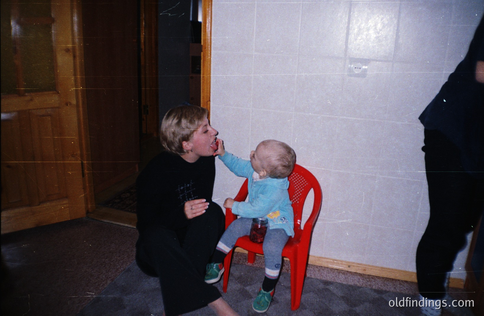 A young girl playfully interacts with a toddler seated in a bright red plastic chair. The scene features textured wallpaper & dark carpeting. Likely a candid snapshot from the 1970s or 1980s, suggesting a family moment. Appears to be indoor residential setting.