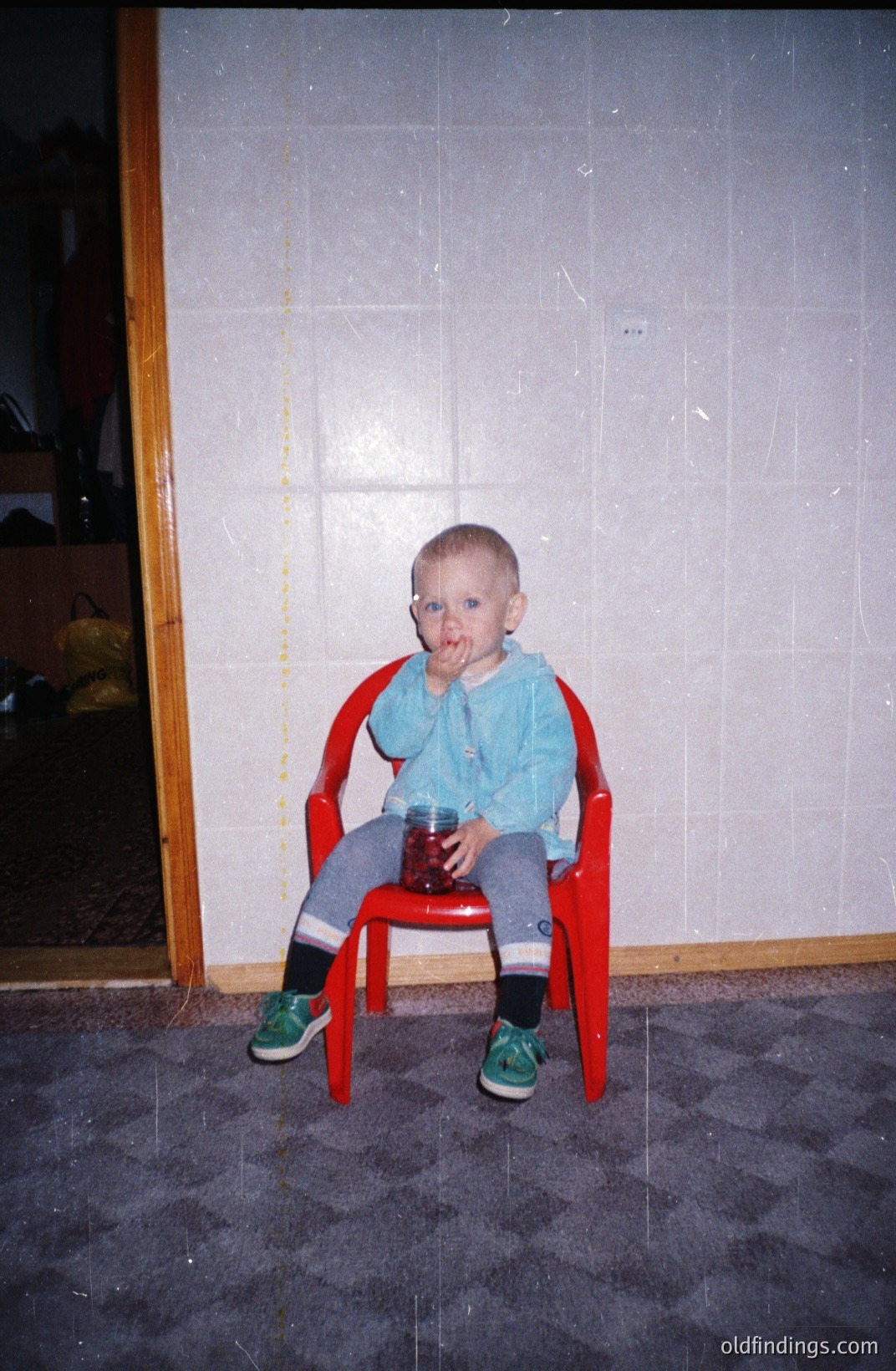 A young child sits in a red plastic chair, covered in red, likely fruit, residue. The child wears blue overalls, striped socks and green shoes. Background features a tiled wall and a glimpse of a darker interior. Likely a family snapshot, circa 1980s.