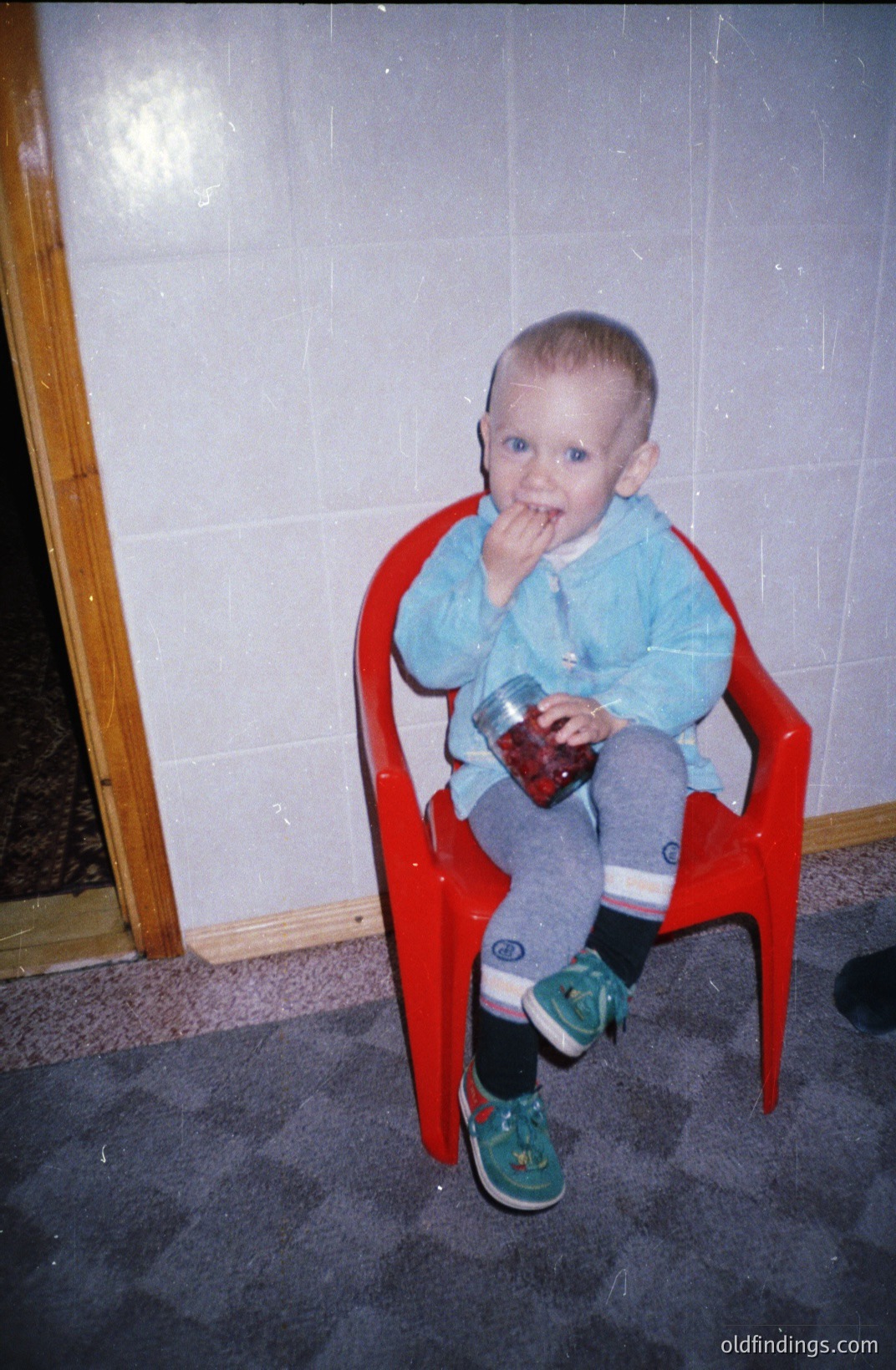 A young child sits in a red plastic chair, intently examining a jar of berries. Visible are green sneakers, dark socks, and a light blue shirt. The backdrop showcases a wall with square tiles and a partial view of a wooden door frame. Likely 1980s home interior. Commercial potential for nostalgic family imagery.