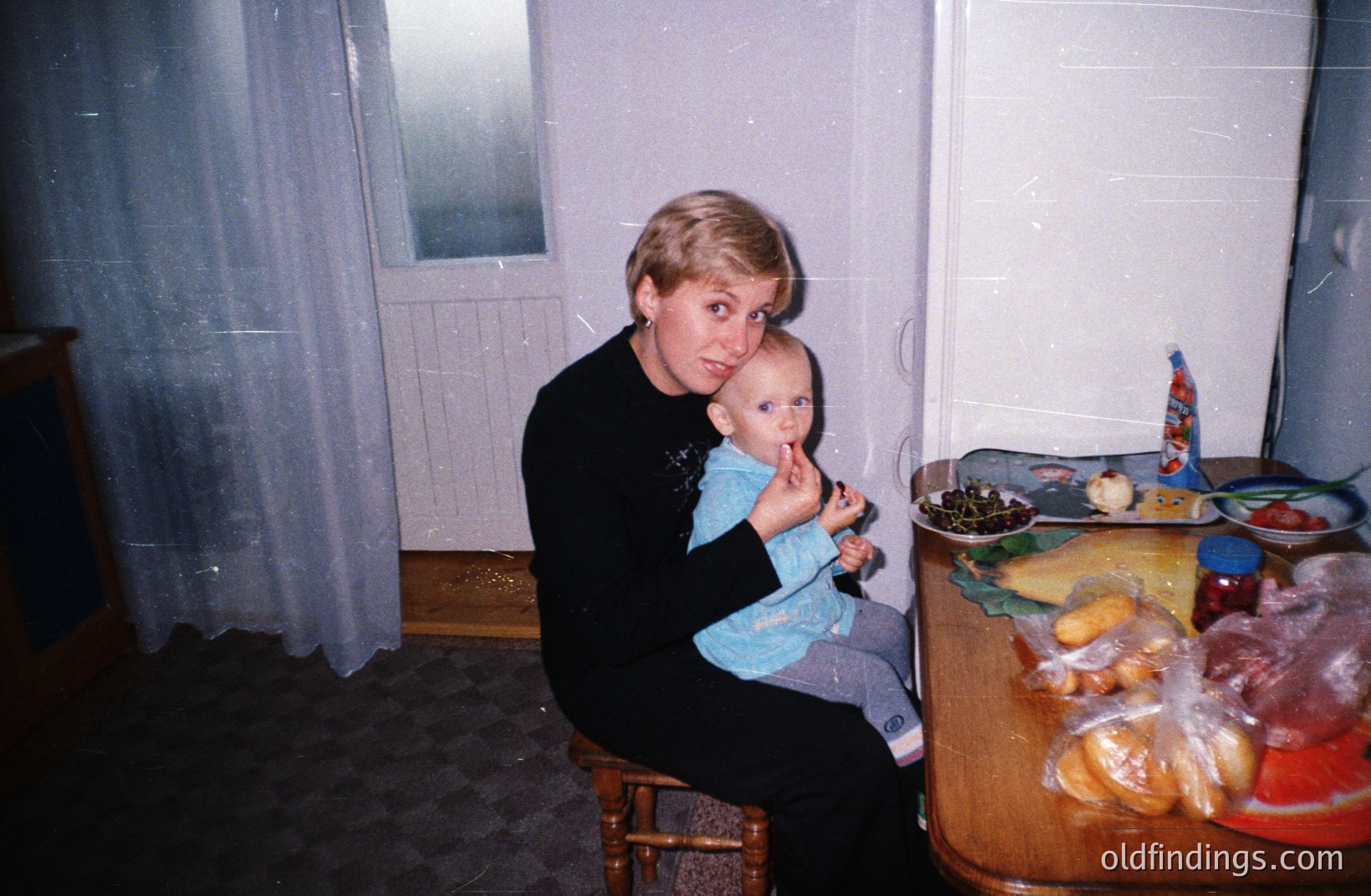 A woman with short blonde hair holds a baby dressed in blue overalls. A richly laden table displays bread, fruit, cheese, and preserved foods. Likely a domestic scene; potential 1980s/90s era based on clothing and photographic style. Simple decor & visible film grain. Could be used for cultural context or design reference.