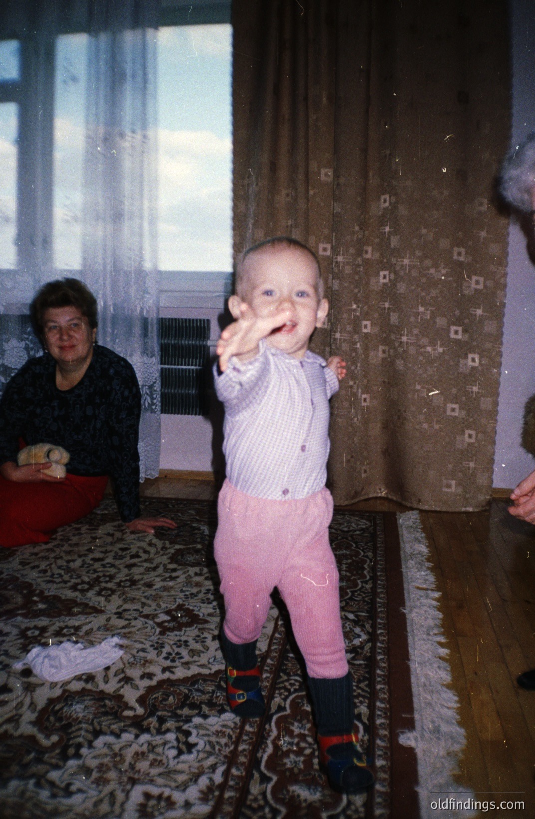 A toddler stands on a patterned rug, raising a hand. Appears to be taking first steps, wearing patterned shirt, pink pants, and dark socks. A woman in a dark patterned dress sits nearby. Likely a family snapshot, mid-1970s style.