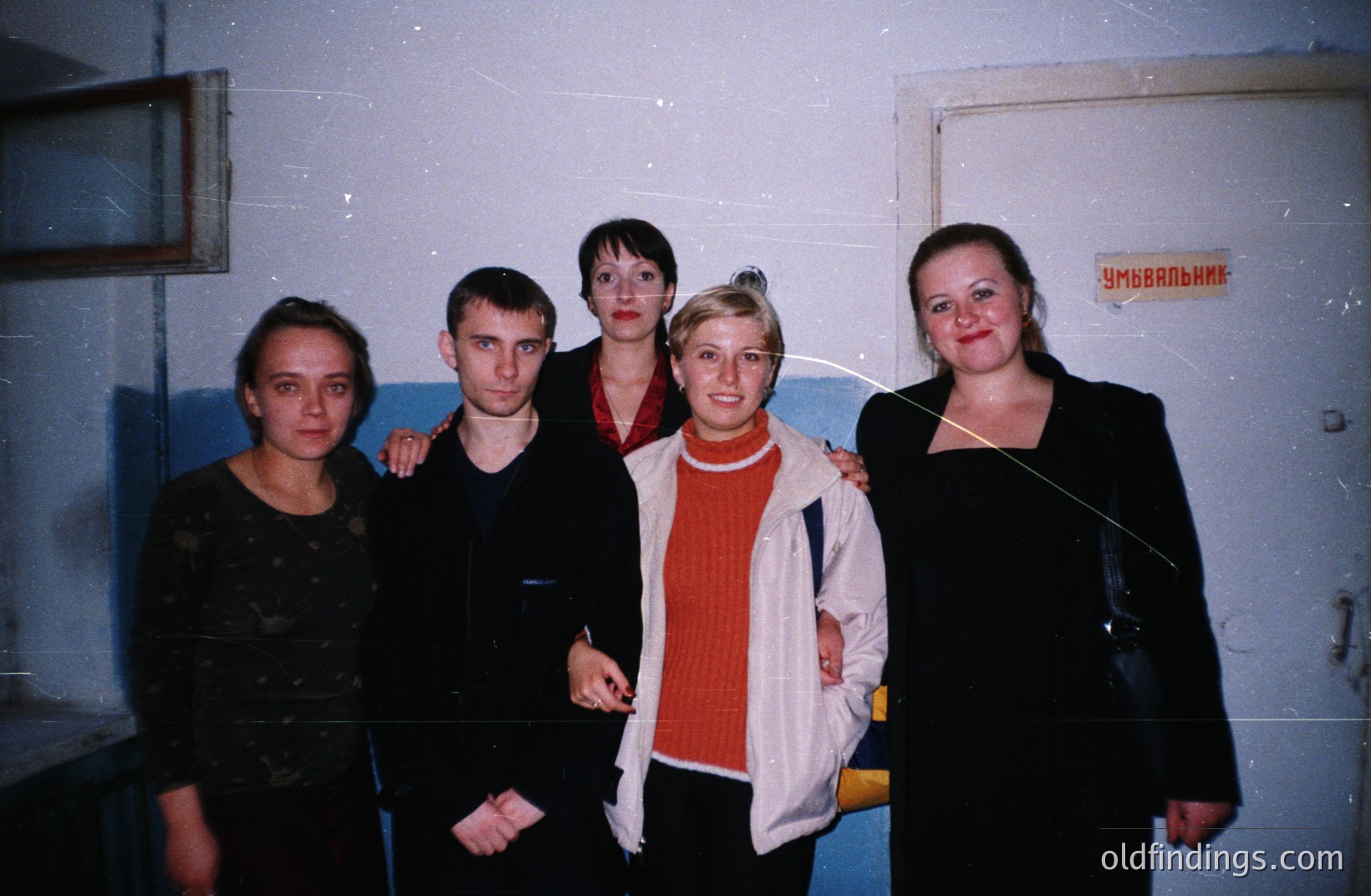 Group portrait of five individuals in a corridor, possibly a public facility. The subjects are dressed in casual attire typical of the 1990s/early 2000s. Cyrillic signage visible above the door suggests a location in Eastern Europe, potentially Bulgaria. Low-key, candid style.
