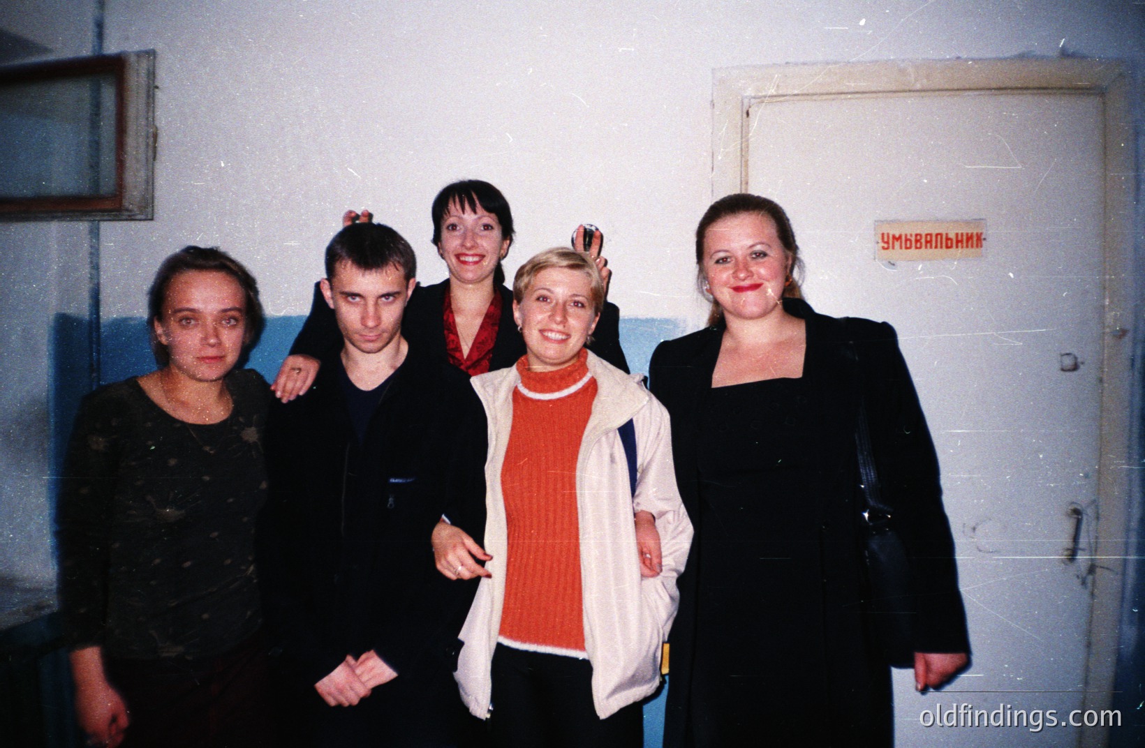 Group portrait of four individuals in a utilitarian space, possibly backstage or a service area. Visible signage in Cyrillic suggests a location in Eastern Europe, potentially Bulgaria. Clothing styles and photography quality indicate a 1990s timeframe. Casual, informal gathering.