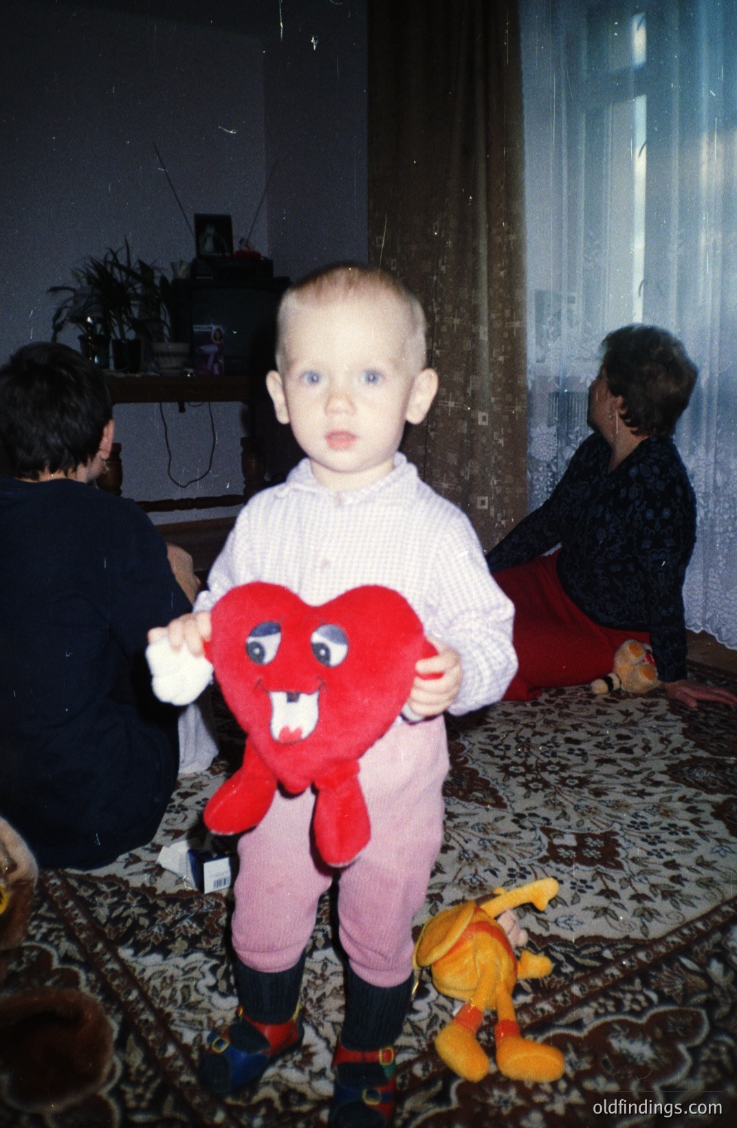 A young boy stands holding a plush, red heart with large eyes and a stitched smile. He wears a patterned shirt & pink pants. A woman sits in the background. Likely a family snapshot, suggestive of the 1970s or 80s. Detailed rug and home decor visible.