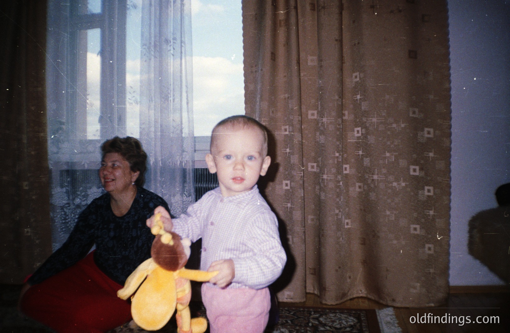 A young child, holding a plush deer, stands indoors with a woman seated in the background. Interior features include patterned curtains and a carpet. Appears to be a snapshot, likely taken in the 1970s. The style suggests family documentation or a personal archive.