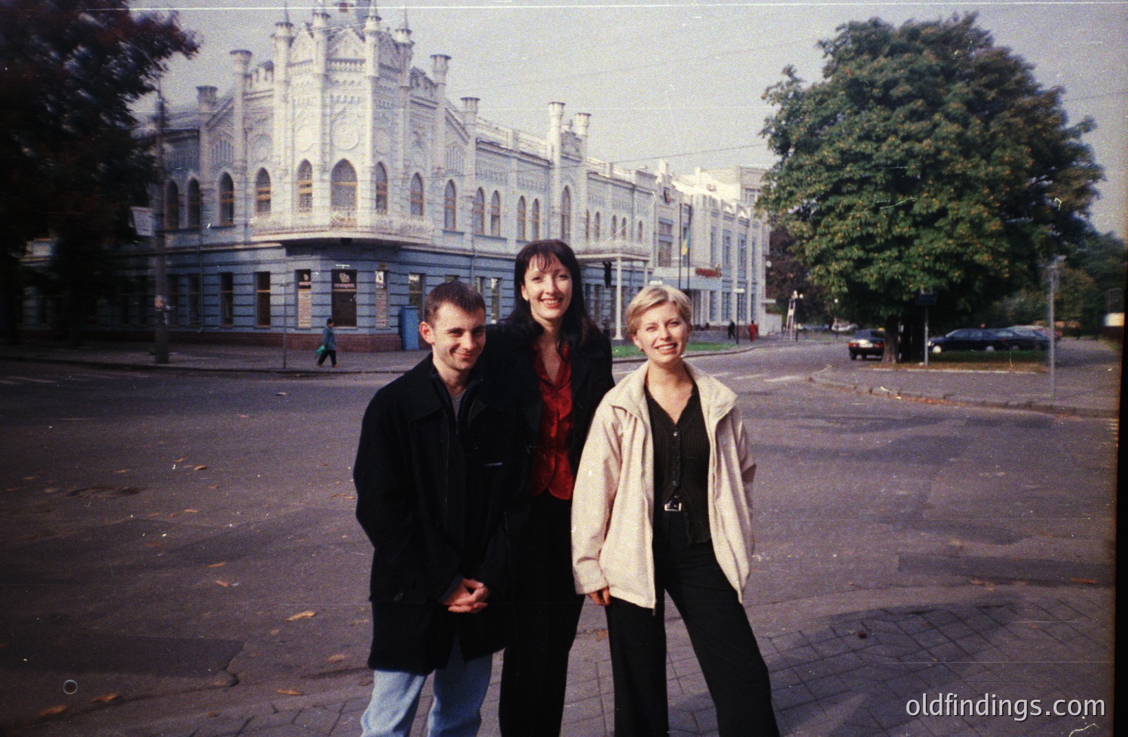 Three people pose in front of a grand, ornate building with numerous arched windows and a central tower, likely government or institutional architecture. The photo, taken on film, exhibits warm tones and a slightly faded aesthetic. Appears to be a city street scene. Possibly Bulgaria, Varna.