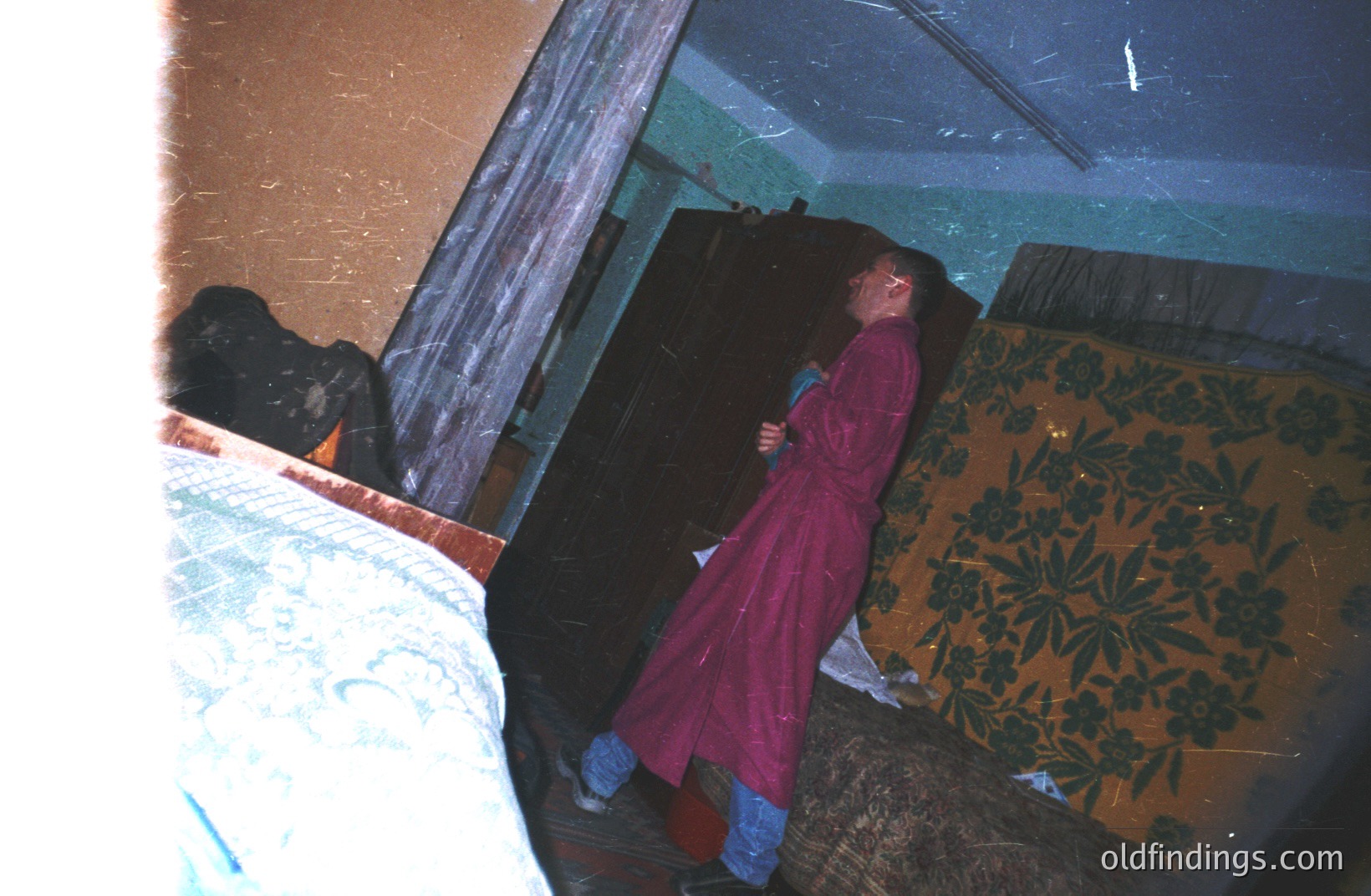 A man in a pink robe and blue jeans stands near a floral-patterned wall, seemingly in a hotel or institutional setting. The room's decor suggests a mid-century aesthetic. A bed frame is visible in the foreground, slightly out of focus. Likely a travel or personal snapshot.