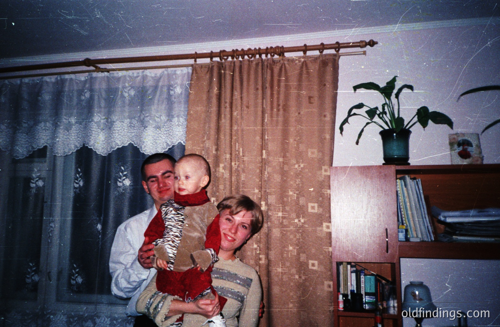 A family portrait: a man, woman, and young child stand near a window with patterned curtains. The child, dressed in red, is held by the man. A large houseplant sits on a bookcase filled with books and objects. Likely 1970s interior style.