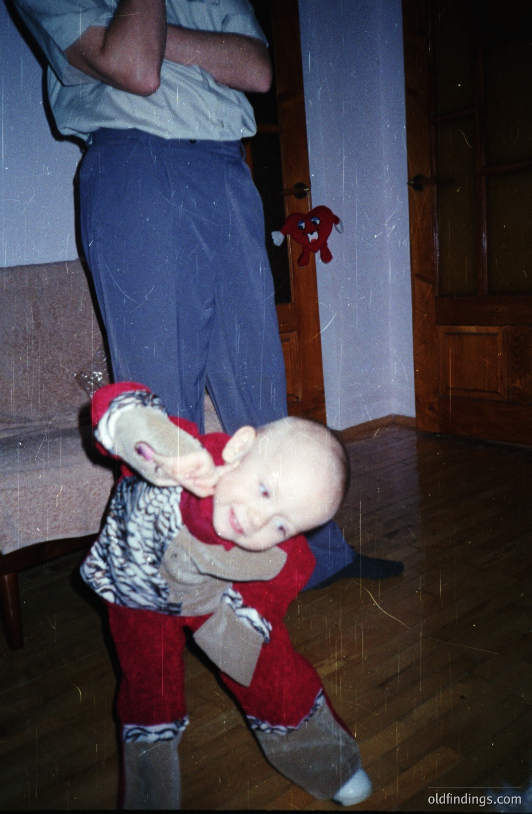A toddler in red and patterned clothing appears to be playfully tipping forward, supported by the legs of an adult. Interior setting with hardwood floors and visible built-in cabinetry. The image displays characteristics of early digital photography—graininess and color cast. Likely a family snapshot, circa 1990s.