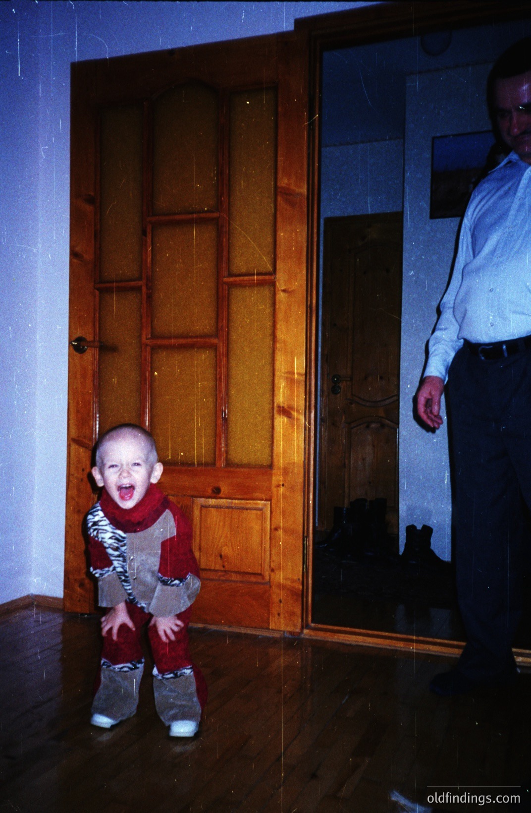 A young child with a distressed expression stands near an ornate, paneled interior door. The child is wearing a patterned jumpsuit. A partial view of an adult in a button-down shirt is visible. Likely a snapshot from the 1970s or 80s, capturing a candid family moment. The image exhibits characteristics of older film stock.